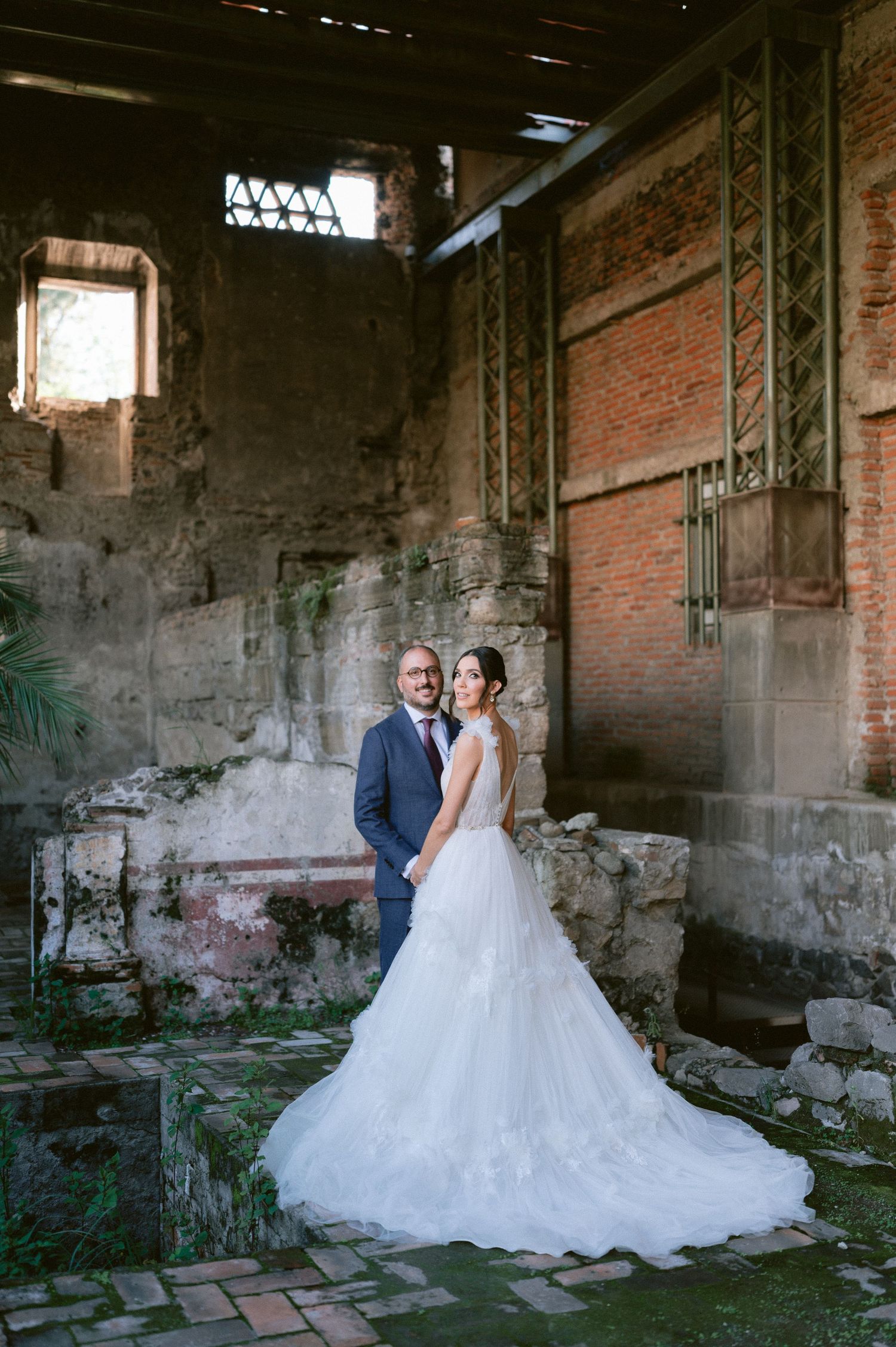 Bride in layered tulle gown and groom in navy suit posing inside weathered brick ruins wedding venue.