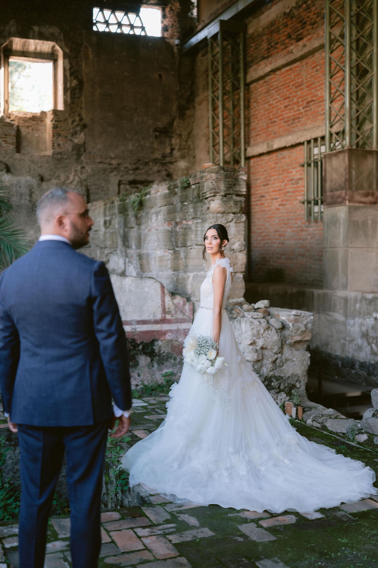 Bride in elegant tiered tulle gown holding bouquet stands before groom inside historic brick ruins.