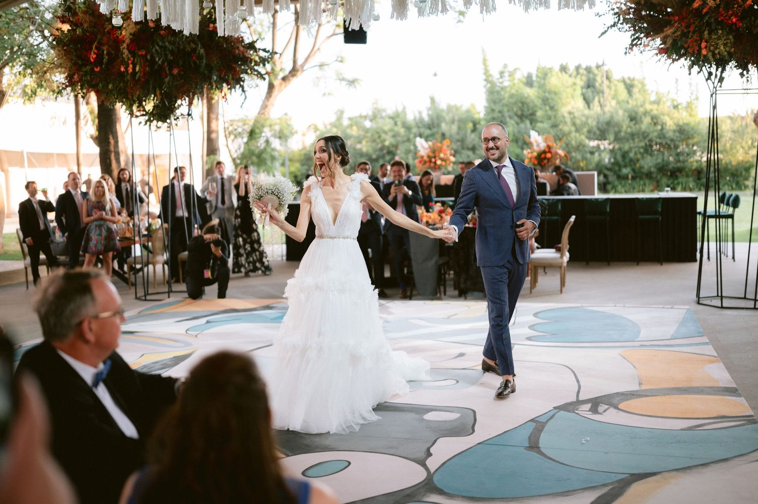 Bride in a layered white gown and groom in navy suit make their grand entrance onto a decorative dance floor.