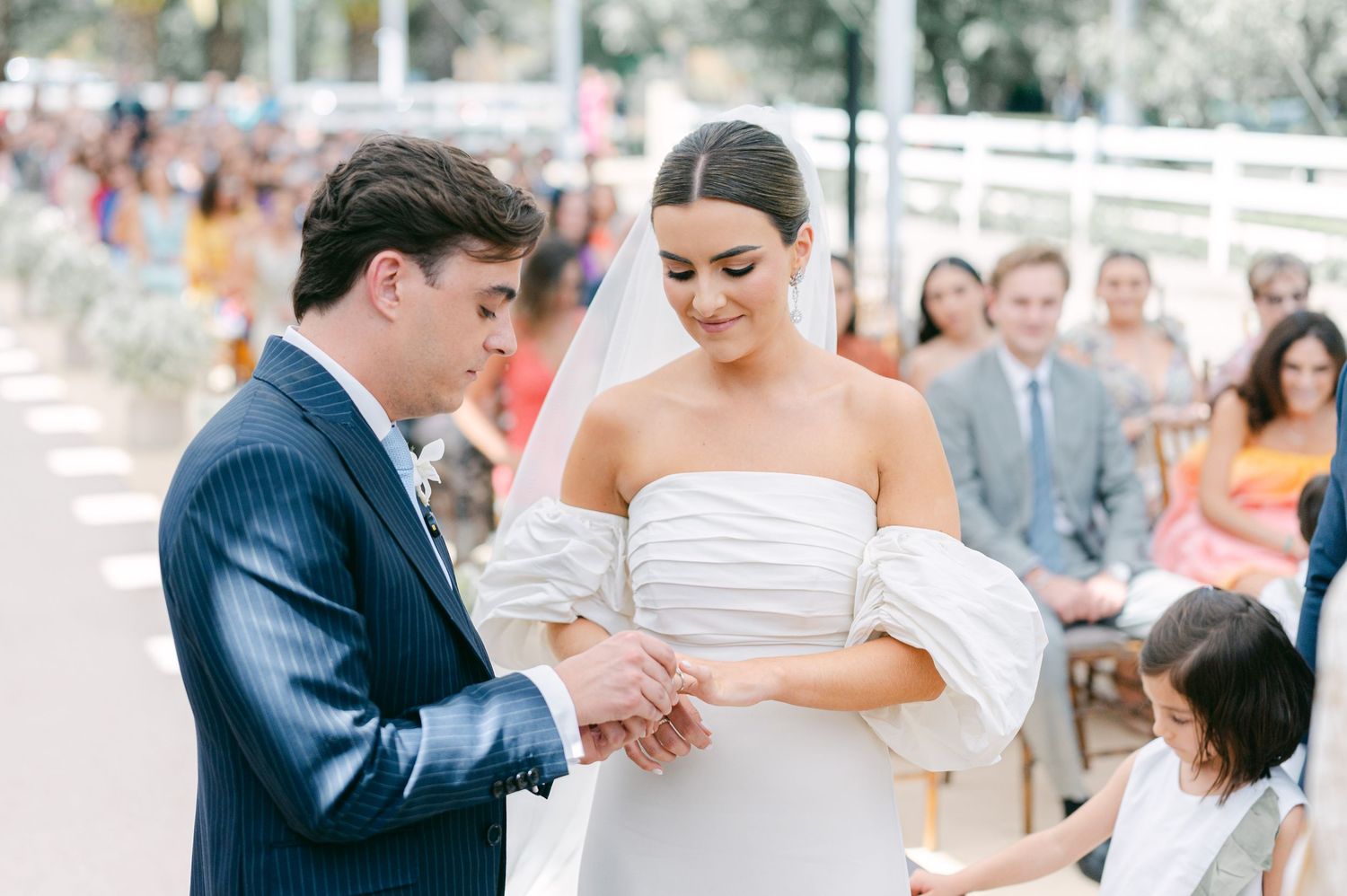 Groom placing wedding ring on brides finger during outdoor ceremony with guests watching.