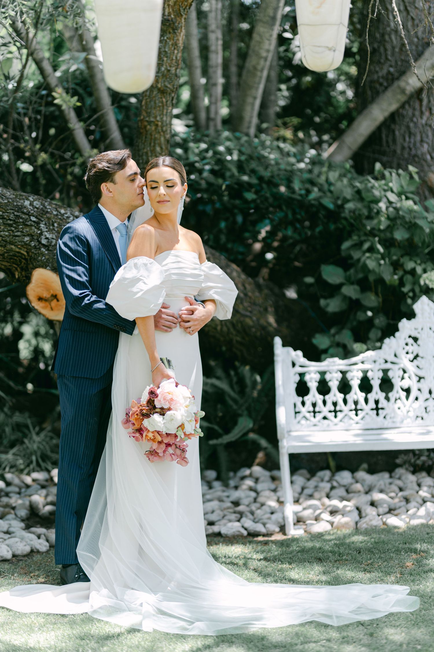 Groom in navy suit embraces bride holding pink floral bouquet near white ornate bench under trees at wedding.