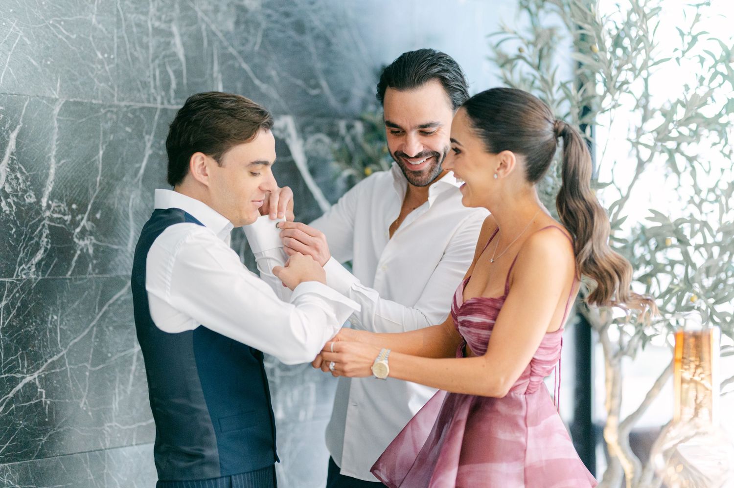 Two men and a woman share a joyful moment adjusting a suit jacket at a wedding preparation.