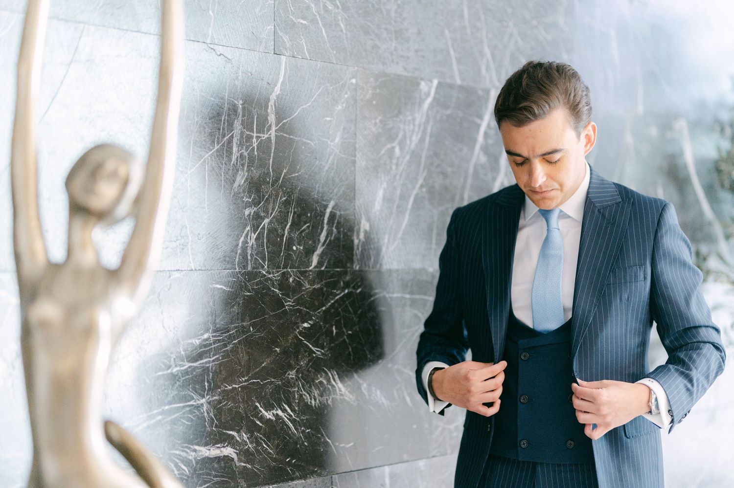 Groom in pinstripe navy suit adjusting jacket buttons beside a bronze statue on marble wall.