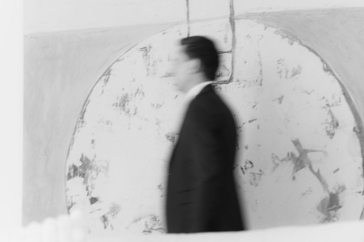 Blurred black and white photo of a man in suit walking past a large circular artwork.