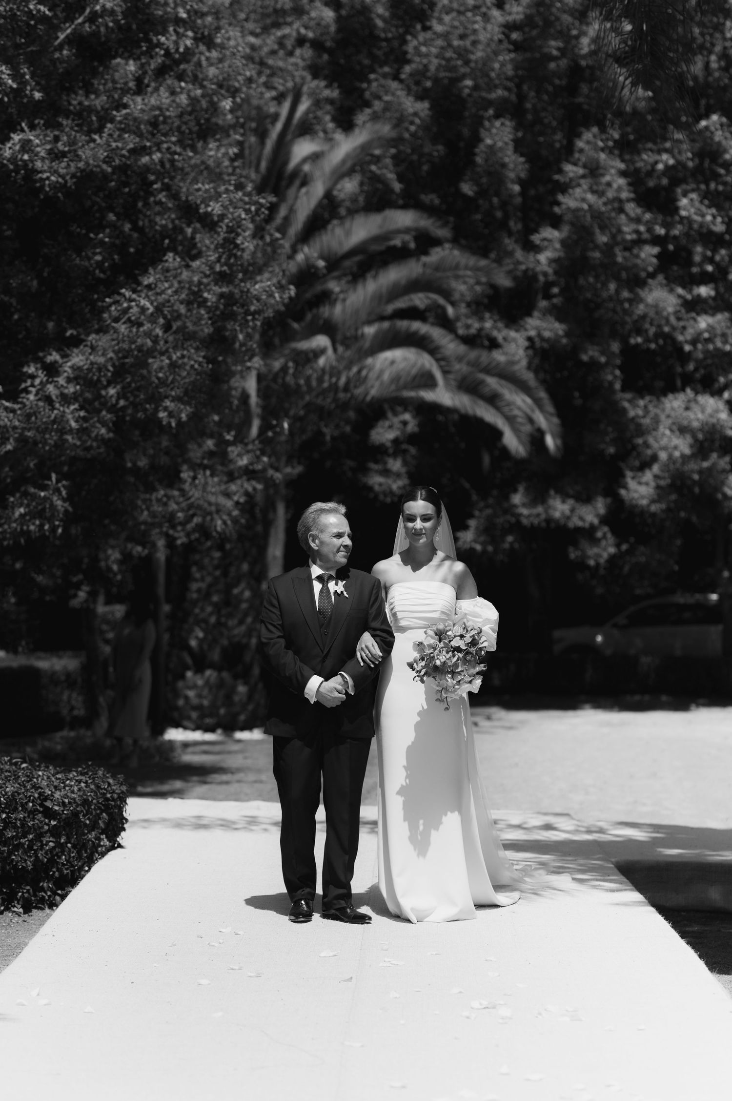 Black and white photo of bride in white gown walking with older man near palm trees.