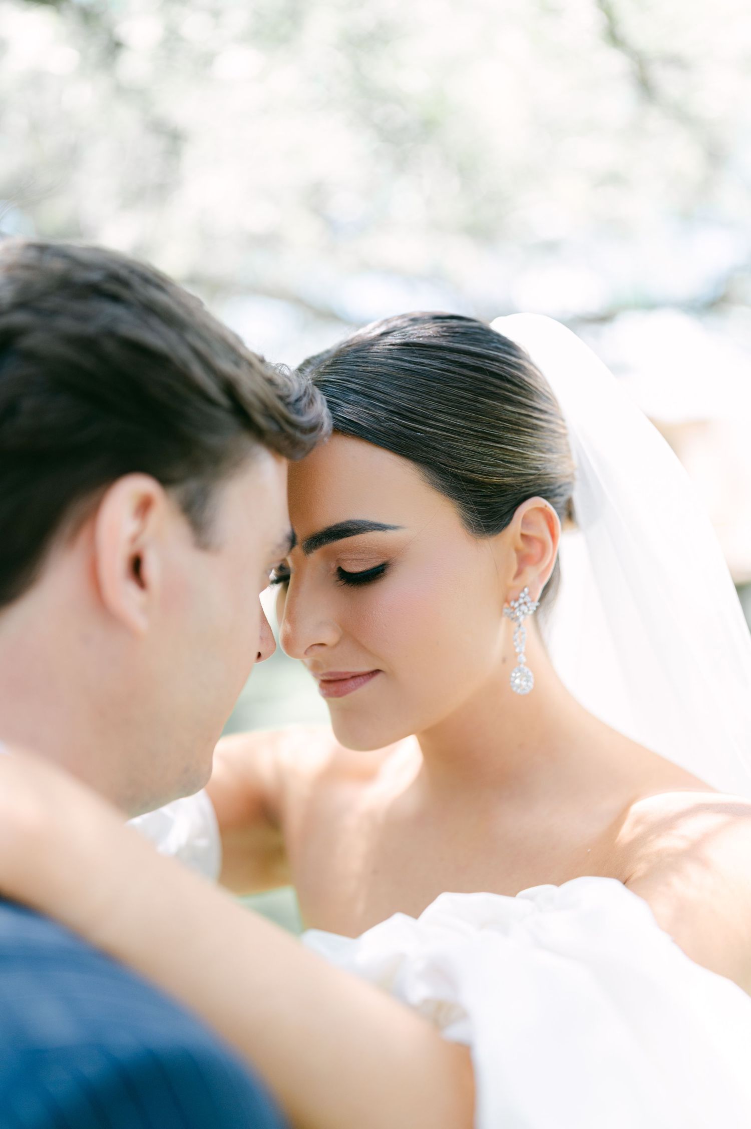 Close-up portrait of bride in veil with elegant crystal earrings leaning toward groom.