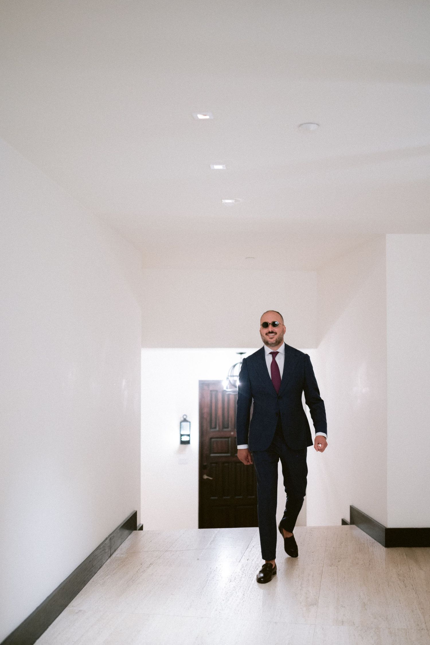 Confident groom in navy suit and burgundy tie walking down bright white hallway on wedding day.