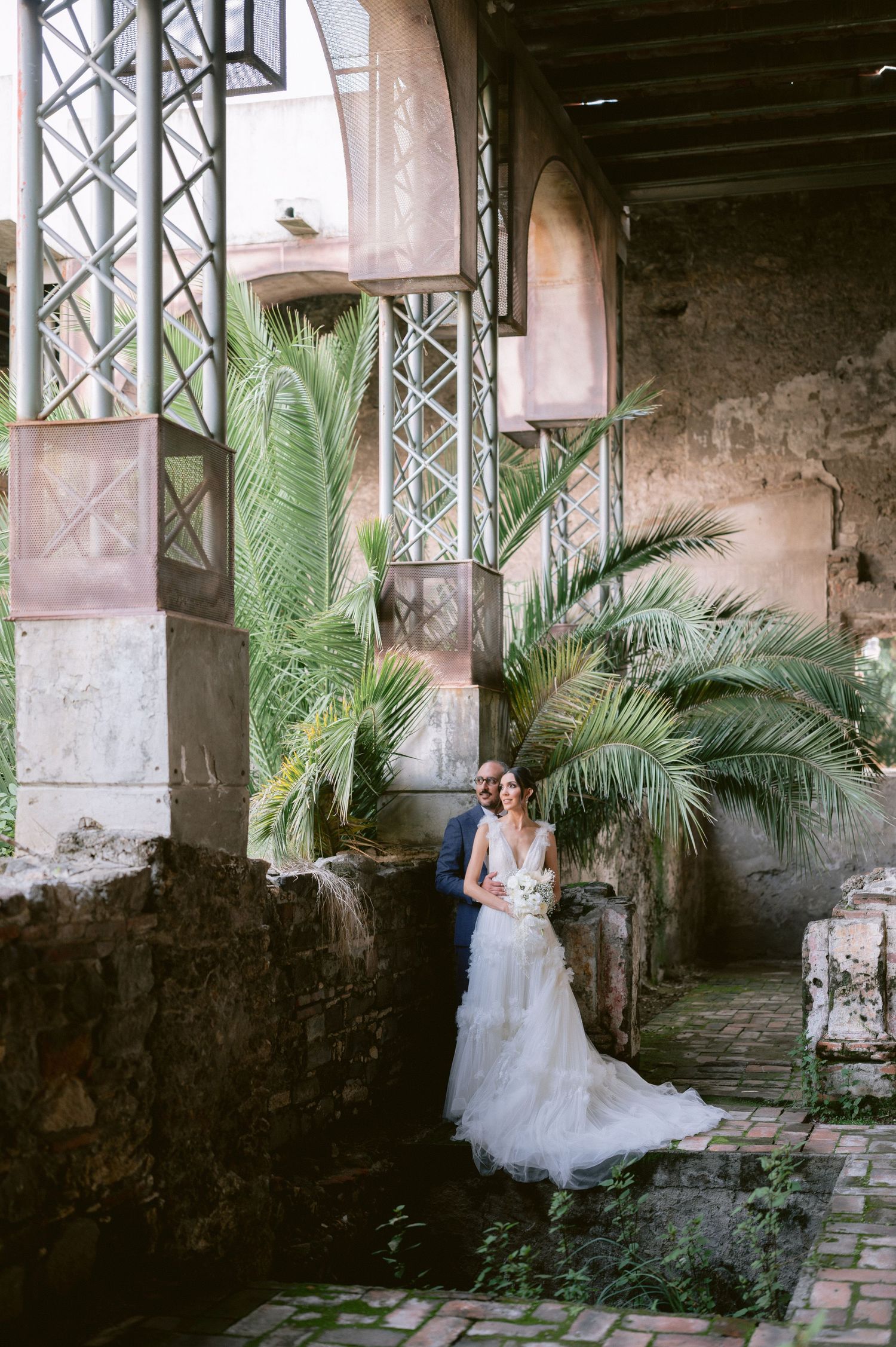 Bride and groom stand romantically beside ornate arched columns with lush tropical palms in historic venue.