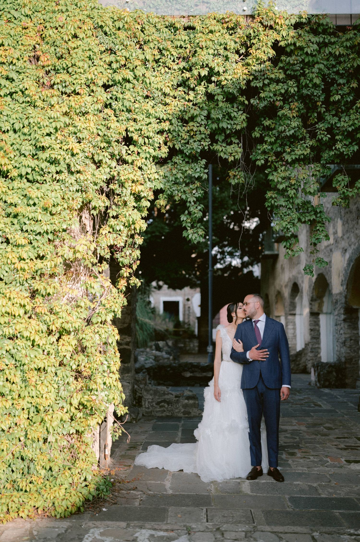 Wedding couple shares a kiss beside lush ivy-covered wall at elegant outdoor venue.