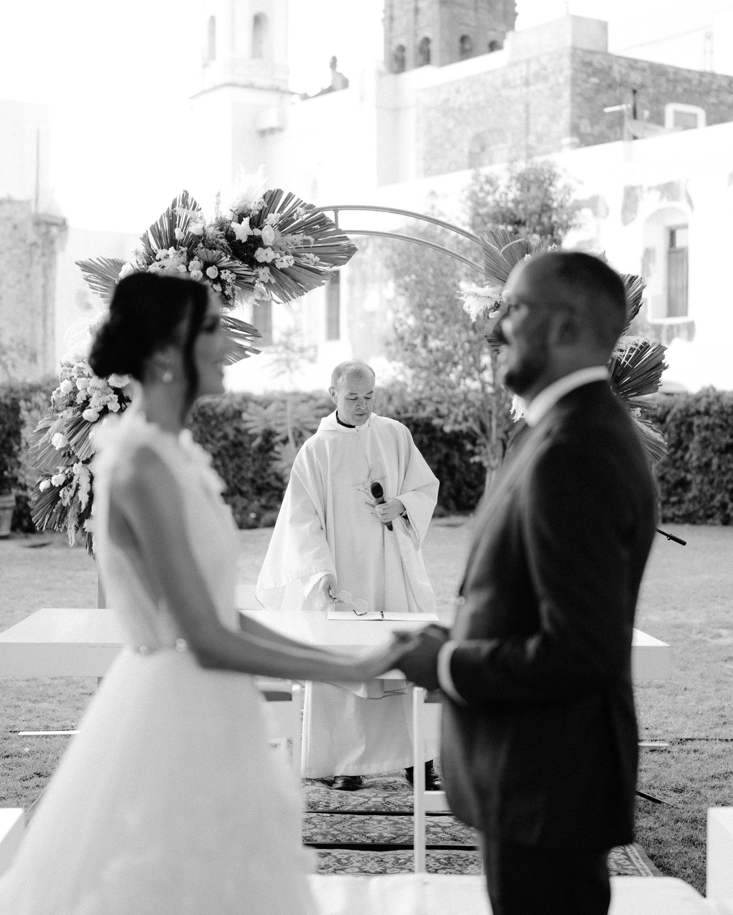 Black and white photo of bride and groom holding hands before priest at outdoor wedding ceremony.