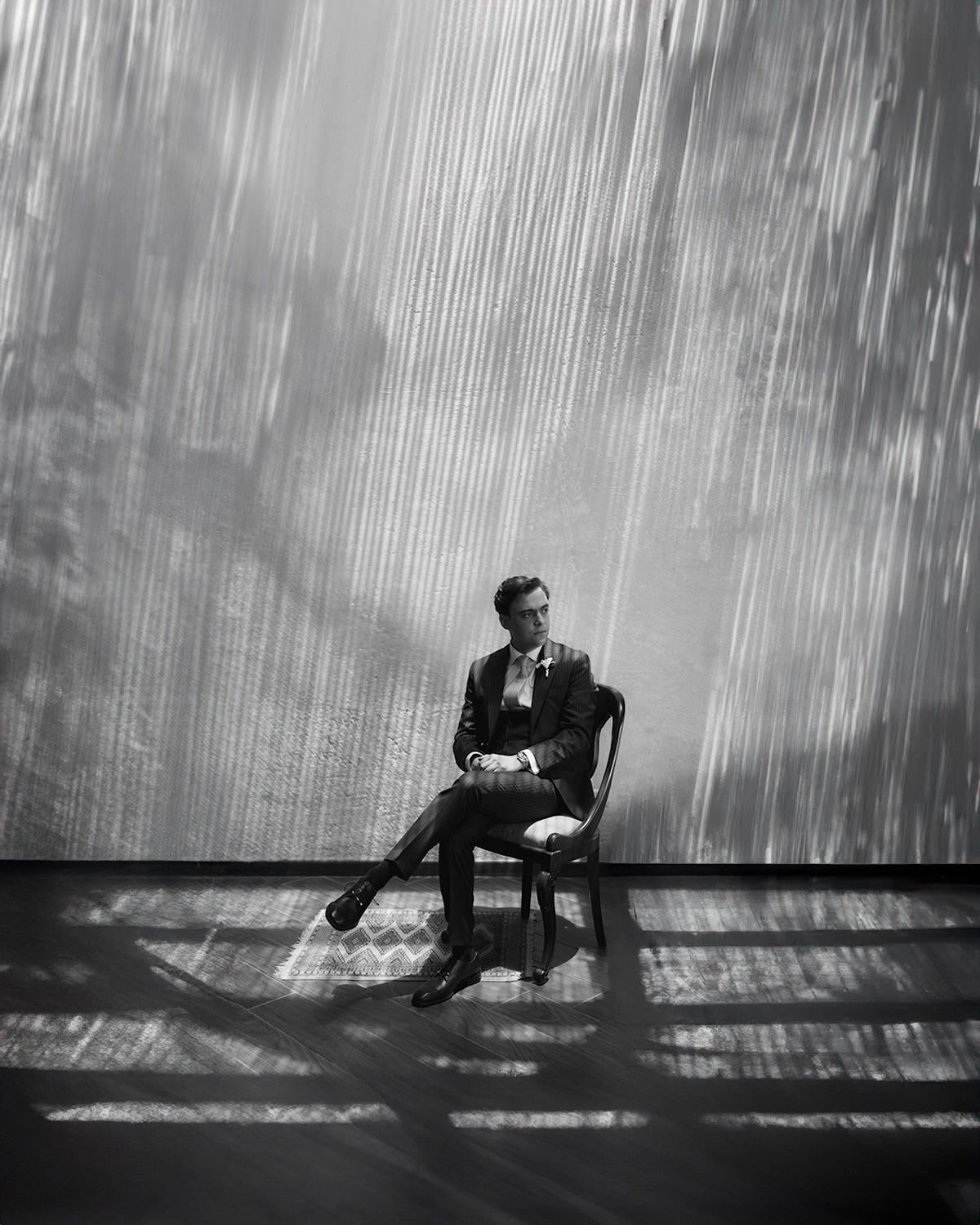 Groom in dark suit seated on chair in dramatic black and white portrait photo.