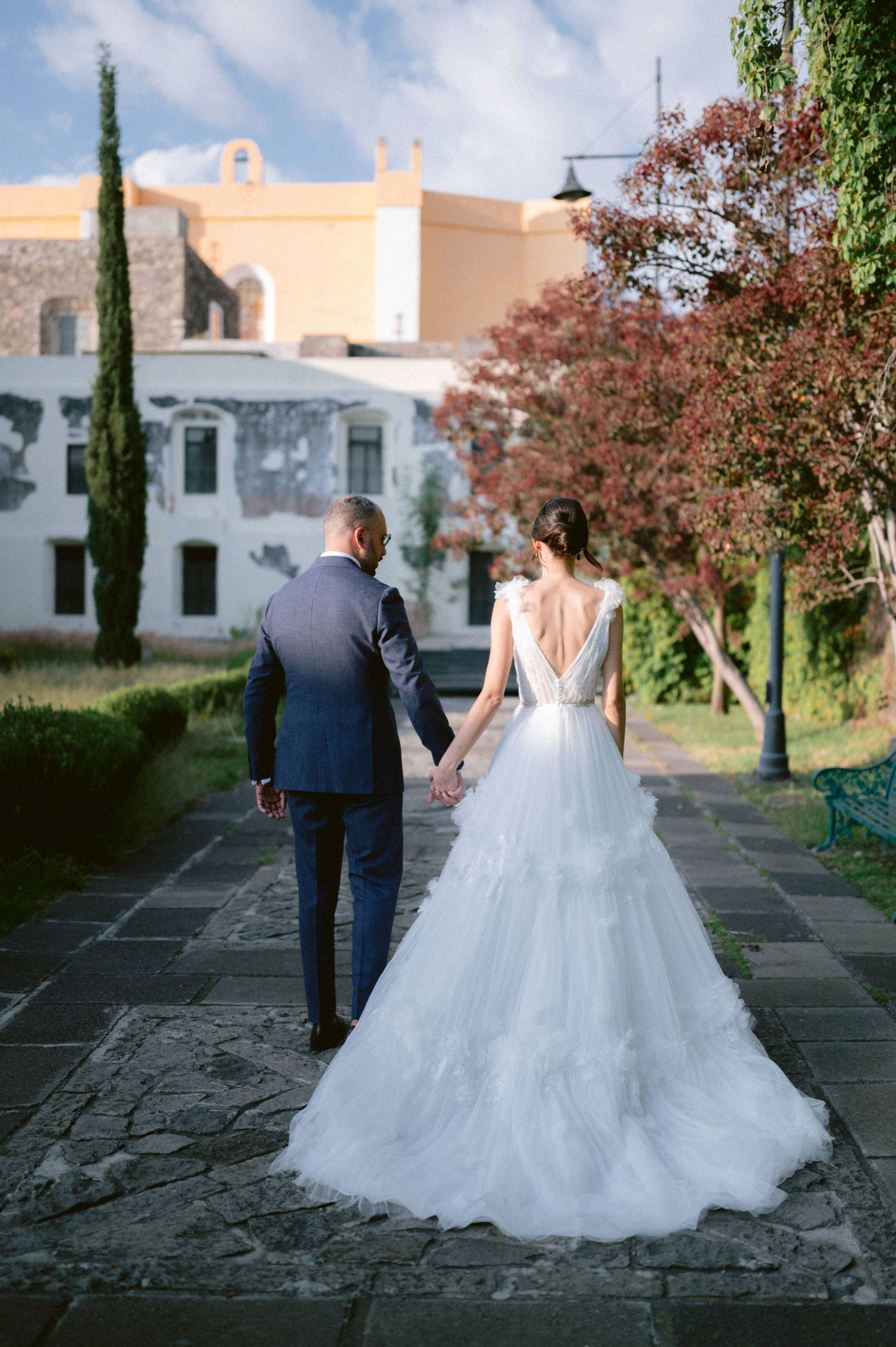 Bride in backless tiered tulle gown holds hands with groom walking through garden venue.