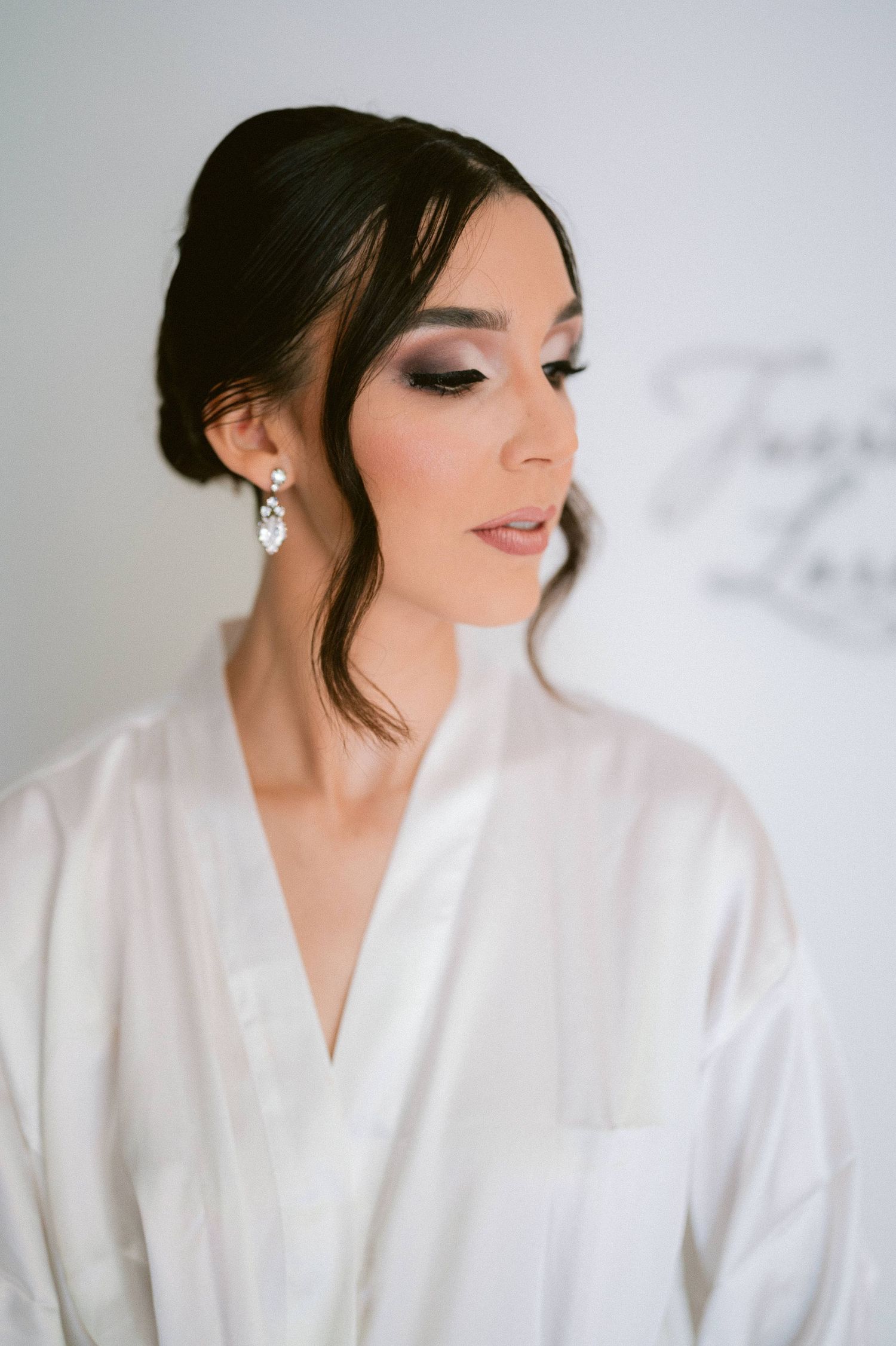 Beautiful bride with dark updo hair, smoky eye makeup, and crystal drop earrings in a white silk robe.