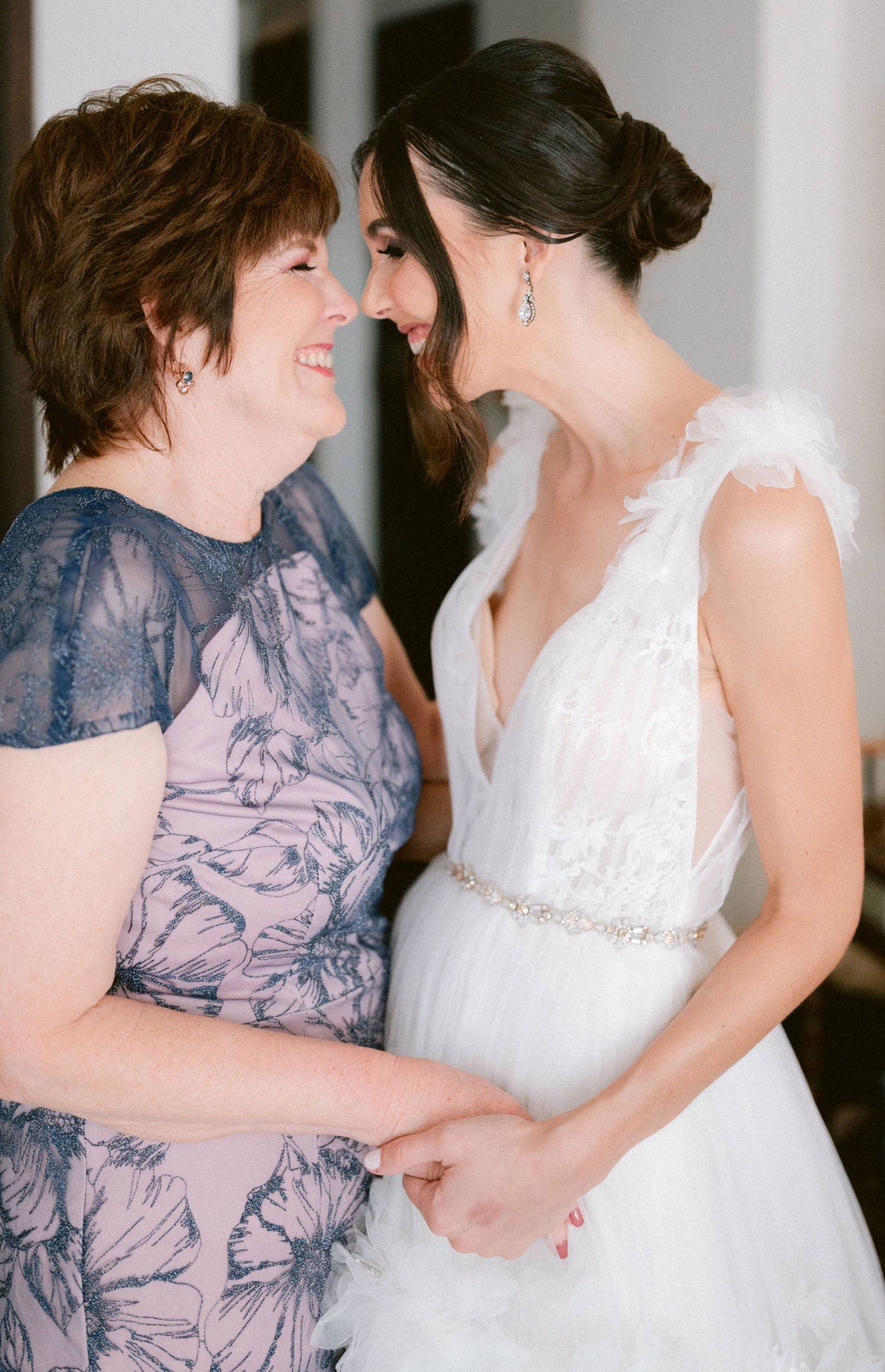 Bride in white feathered gown shares tender moment with mother in floral dress on wedding day.