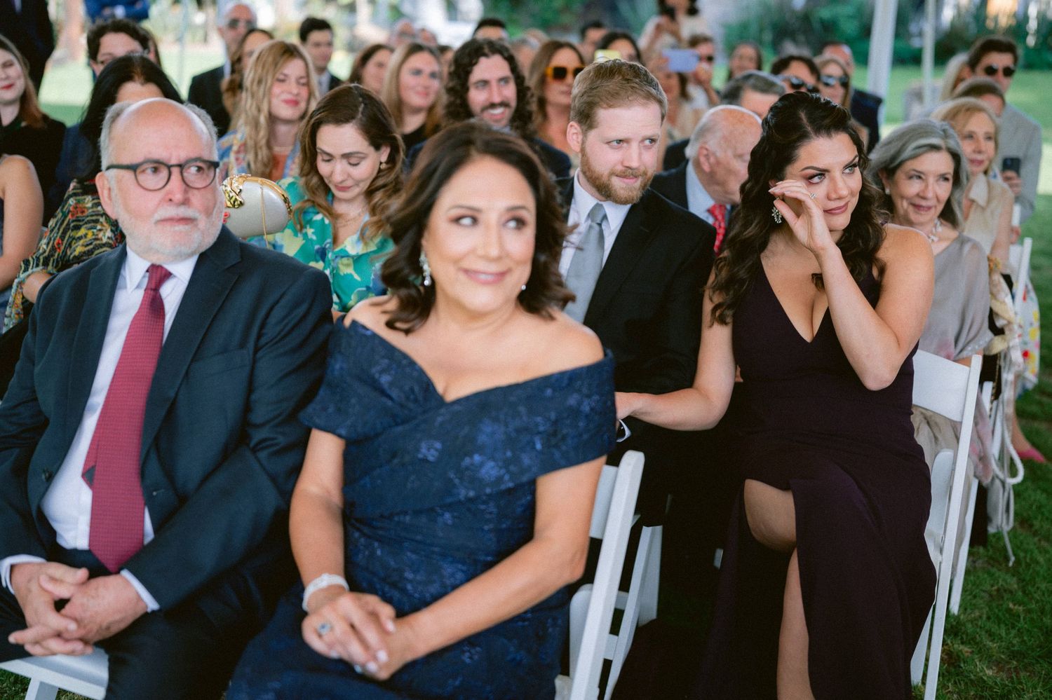 Wedding guests in formal attire seated at outdoor ceremony, woman in navy off-shoulder dress smiling.