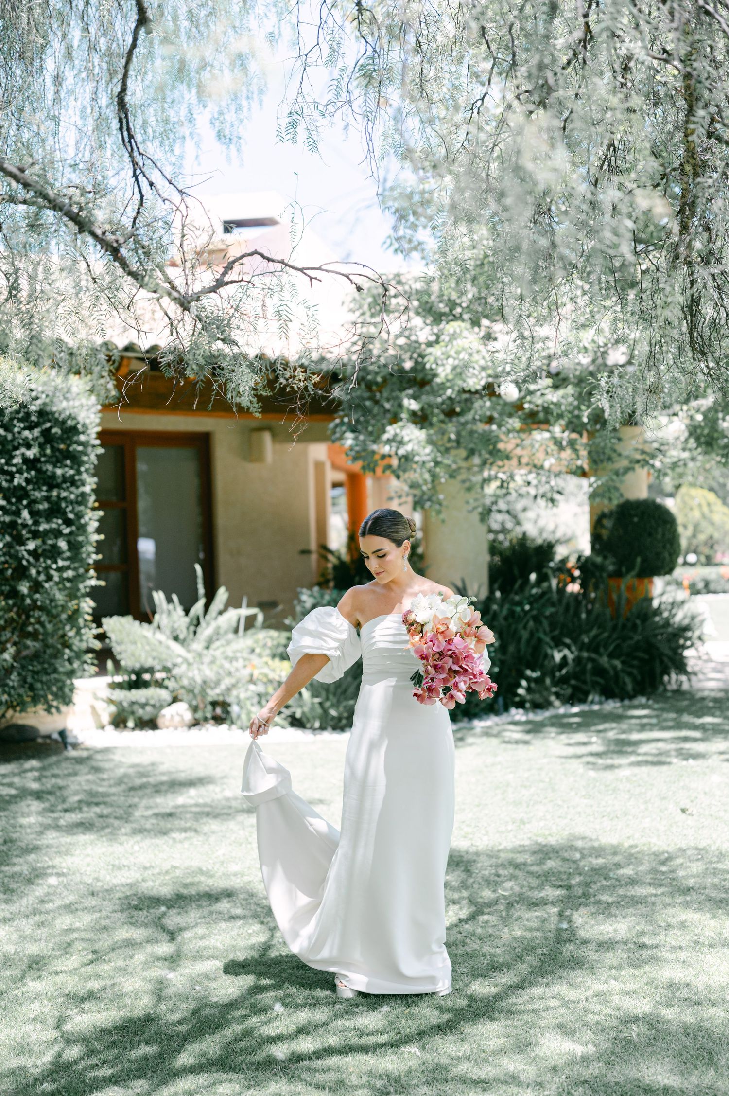 Bride in off-shoulder white gown twirls dress while holding pink orchid bouquet in lush garden setting.