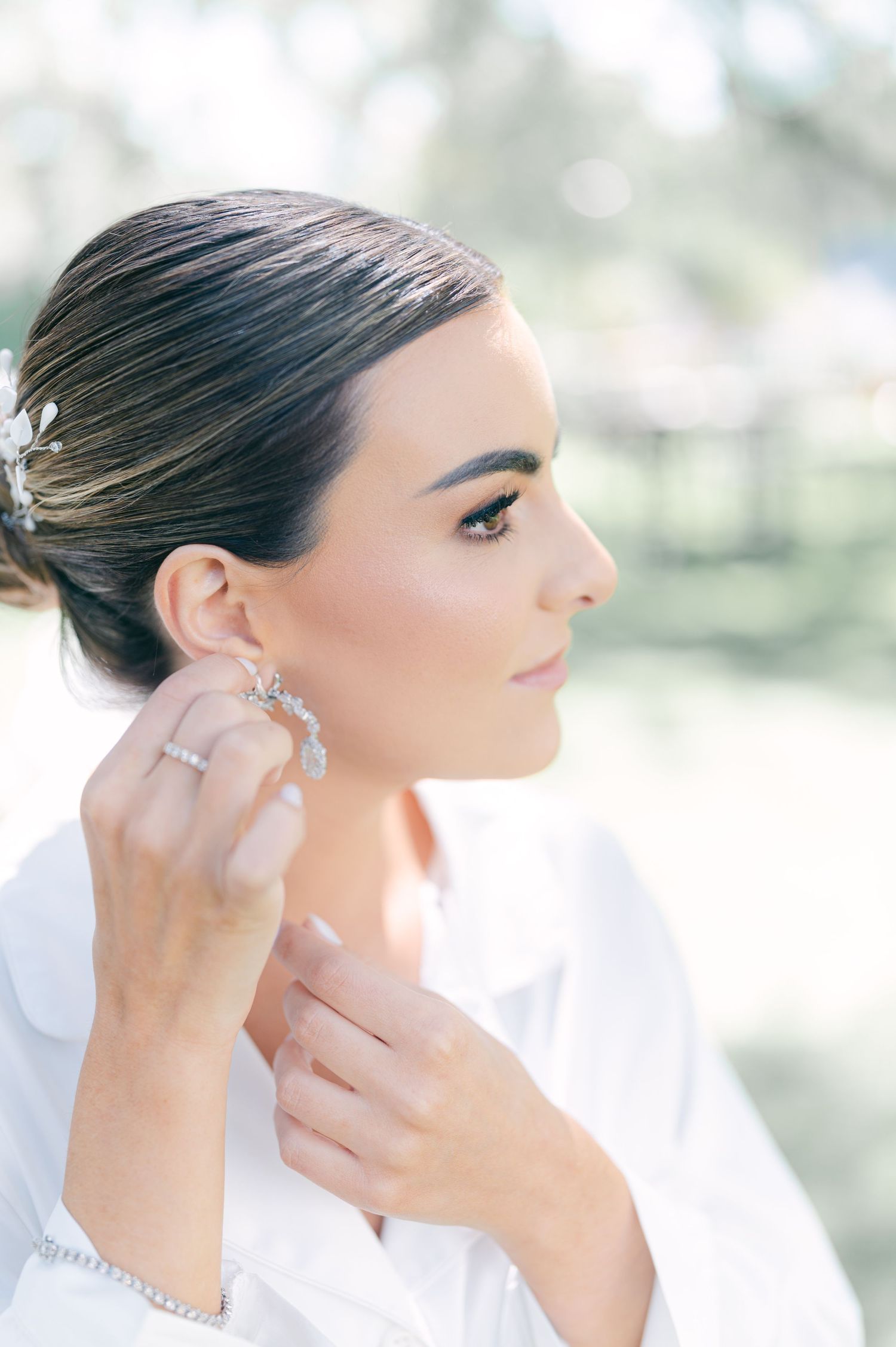 Bride in white robe putting on elegant crystal drop earrings with hair in sleek updo outdoors.