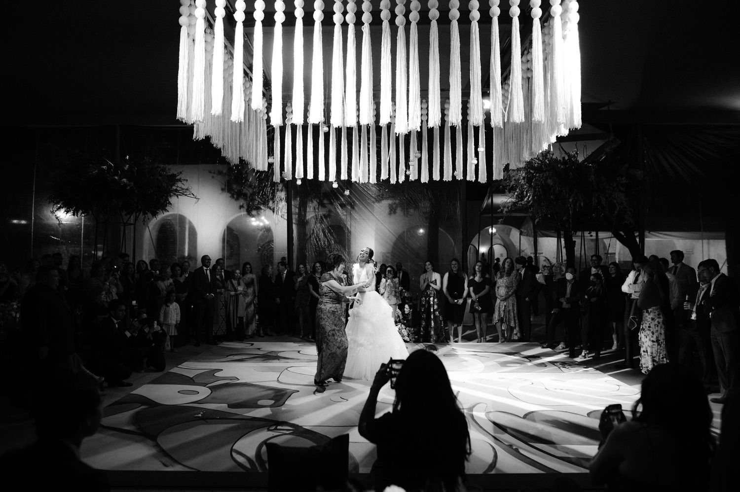 Black and white wide shot of bride and groom first dance under glowing chandelier with guests watching.