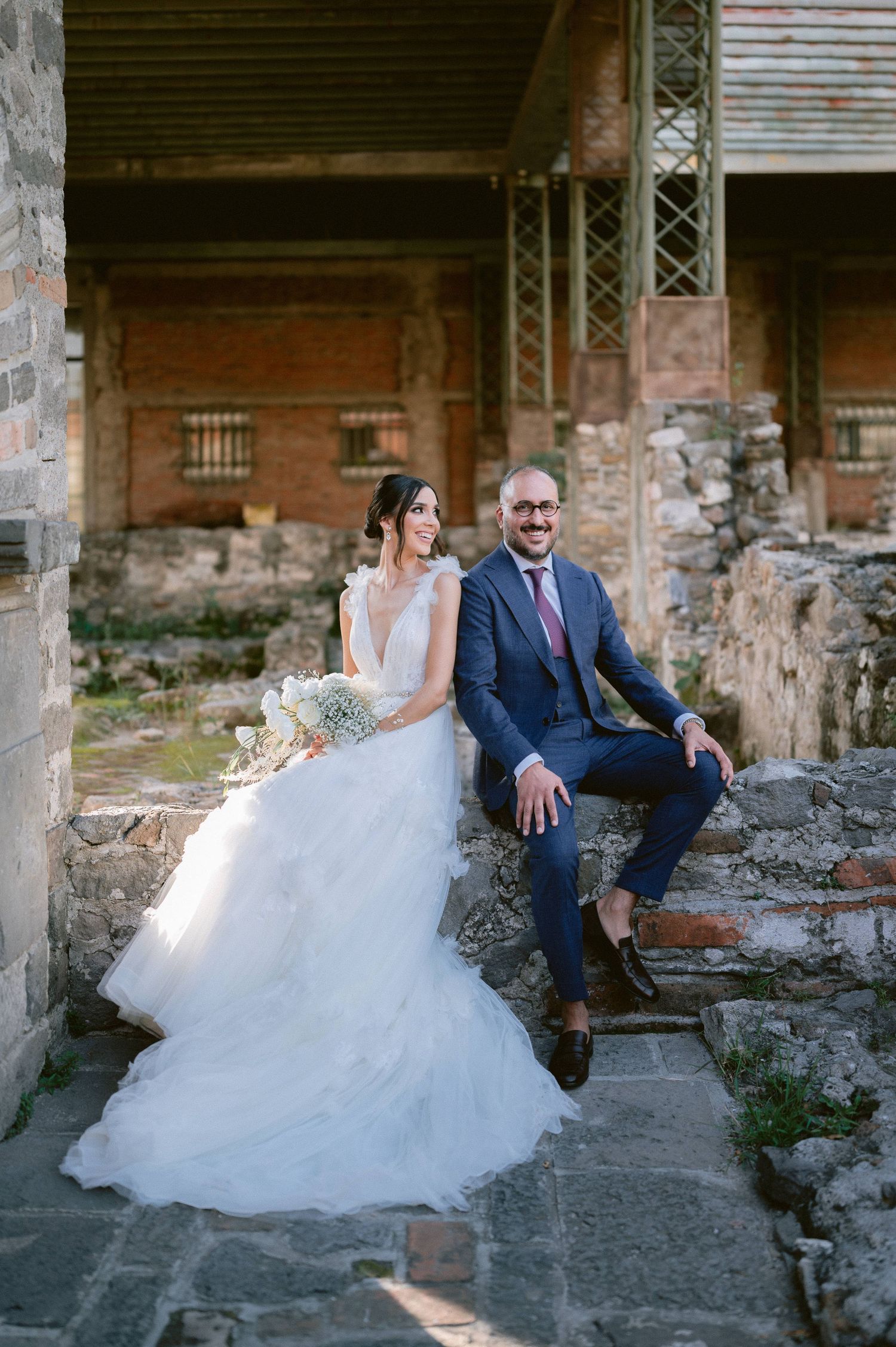 Bride in white tulle gown and groom in navy suit sitting on stone steps at rustic ruins wedding venue.