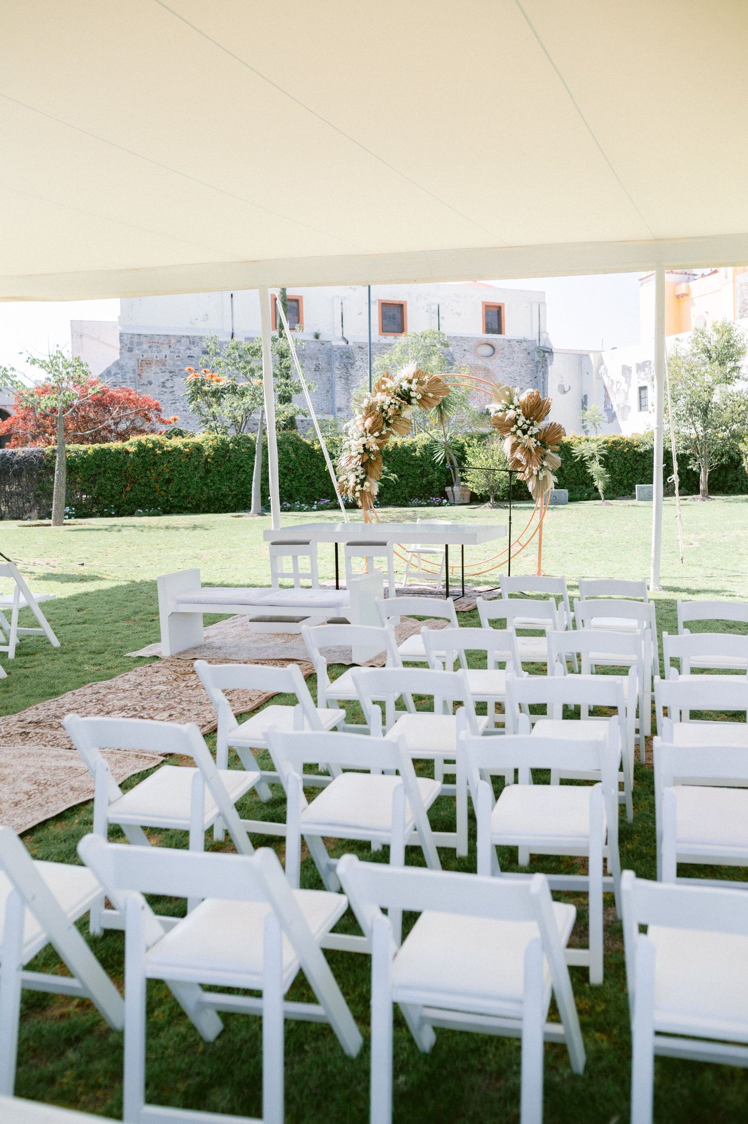 Outdoor wedding ceremony setup with white folding chairs under a tent facing a floral arch.