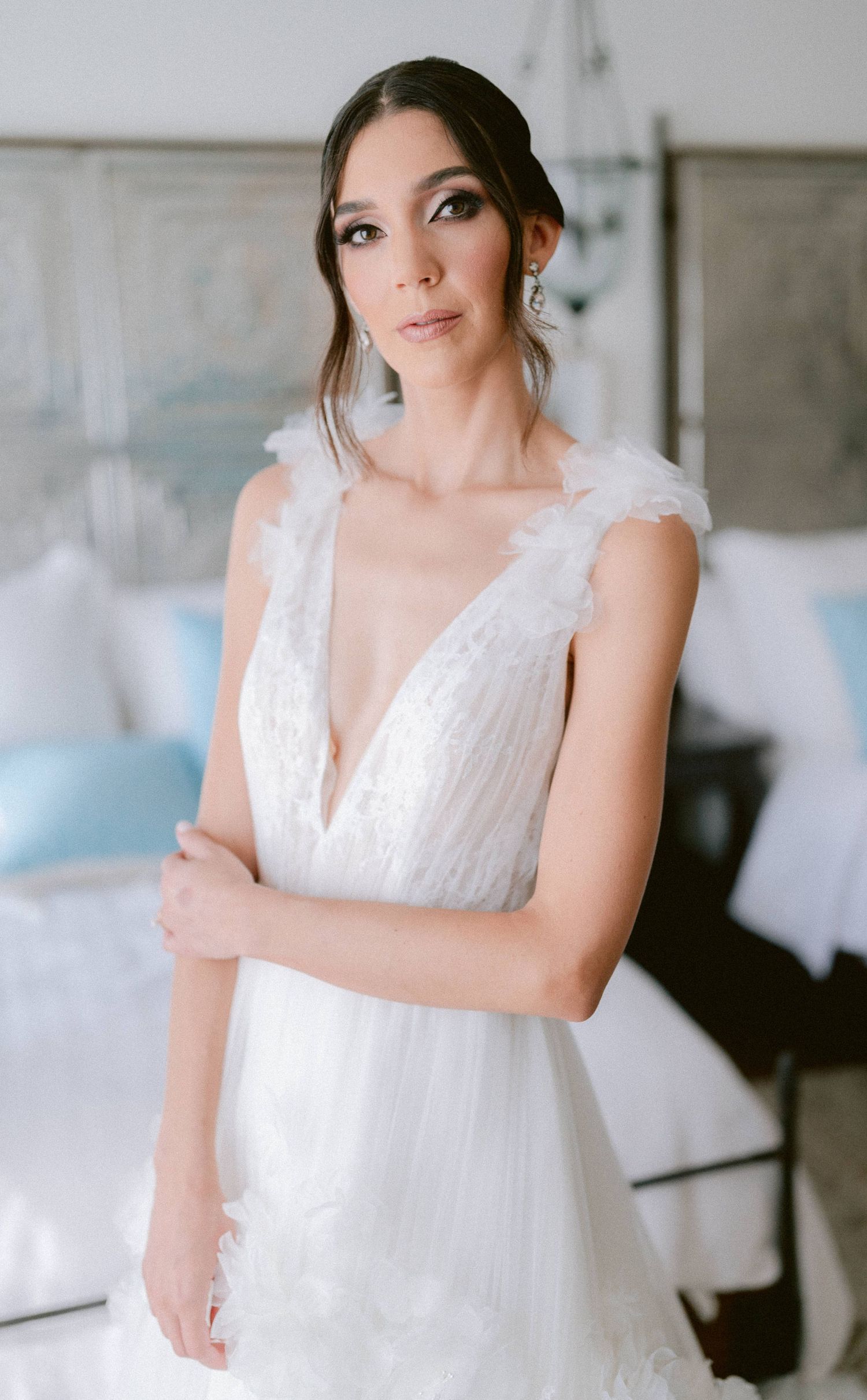Elegant bride in deep V-neck feather-trimmed wedding dress poses with arms crossed indoors.