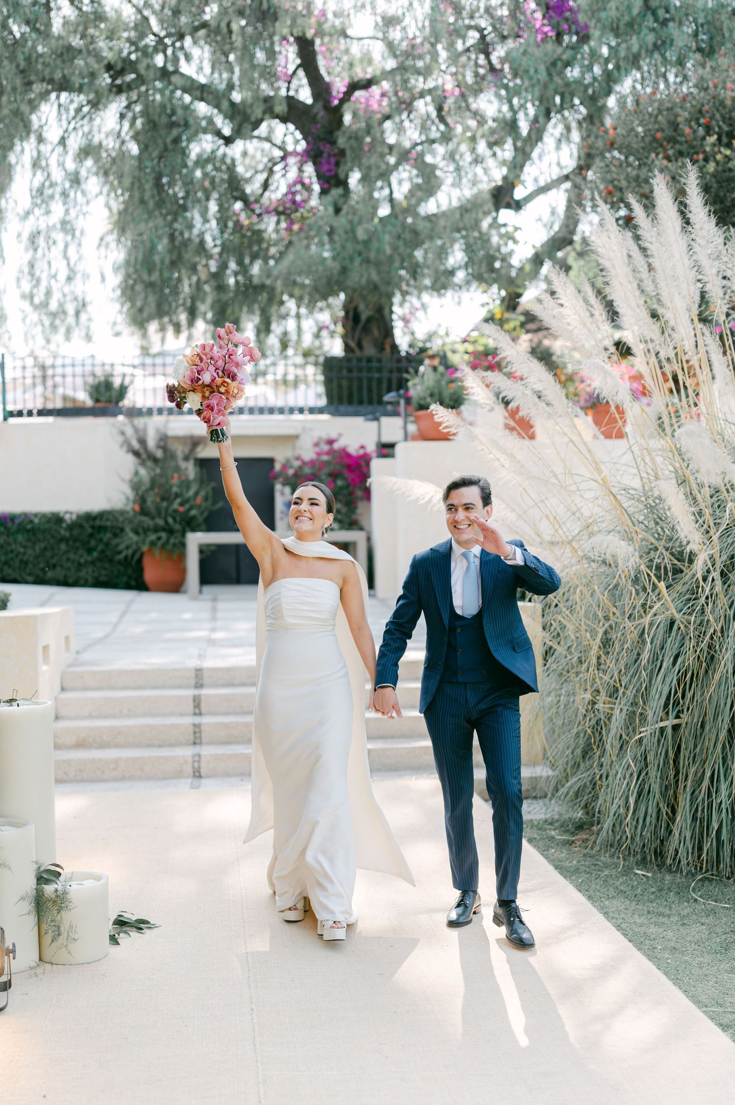 Celebrating newlyweds walk hand in hand as bride raises colorful bouquet in garden setting.
