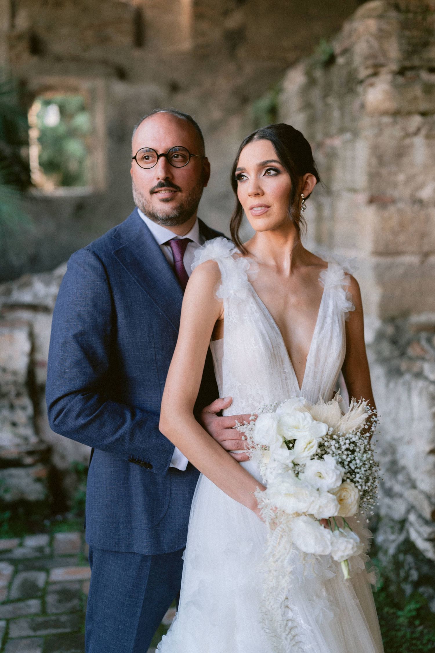 Bride and groom pose together at ancient stone ruins, she holds white bouquet in deep V-neck tulle gown.