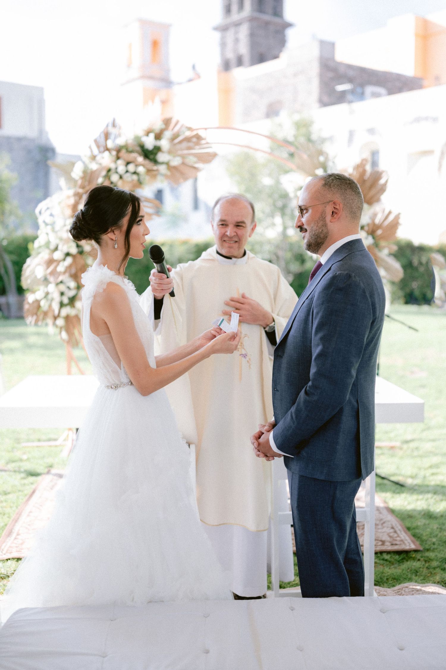 Bride reads vows to groom while smiling officiant holds microphone at sunny outdoor wedding ceremony.