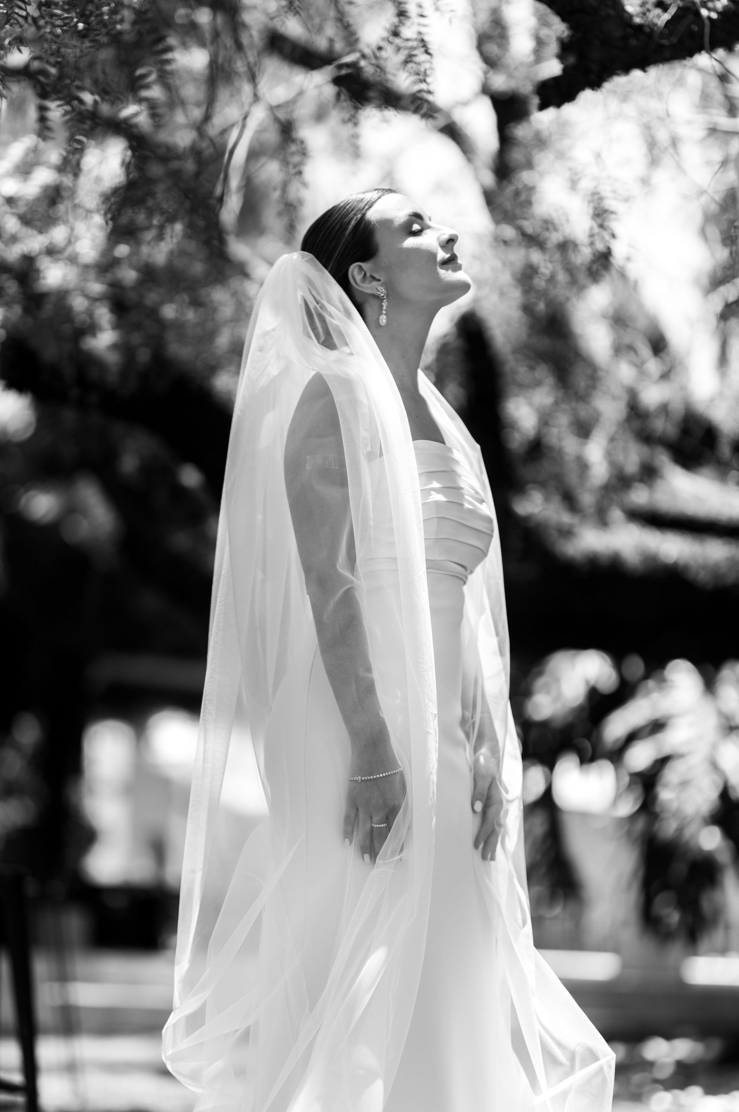 Elegant bride in white gown and veil gazes upward outdoors in black and white wedding photo.