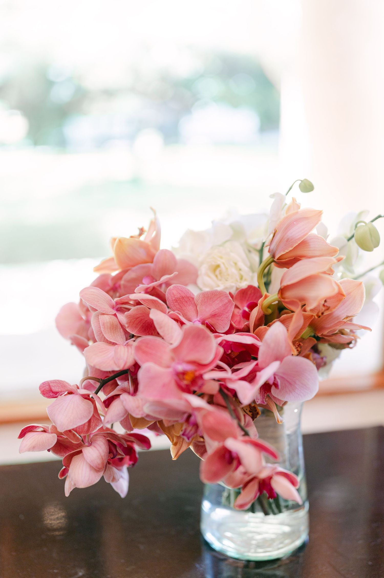 Romantic bridal bouquet of pink orchids and white roses arranged in a clear glass vase by window.