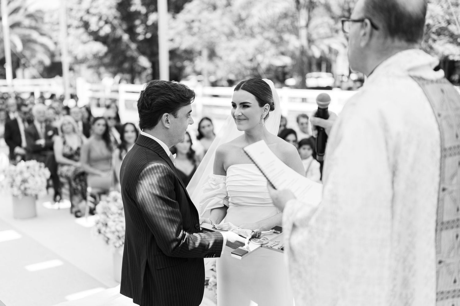 Black and white photo of bride and groom exchanging vows with officiant at outdoor wedding ceremony.