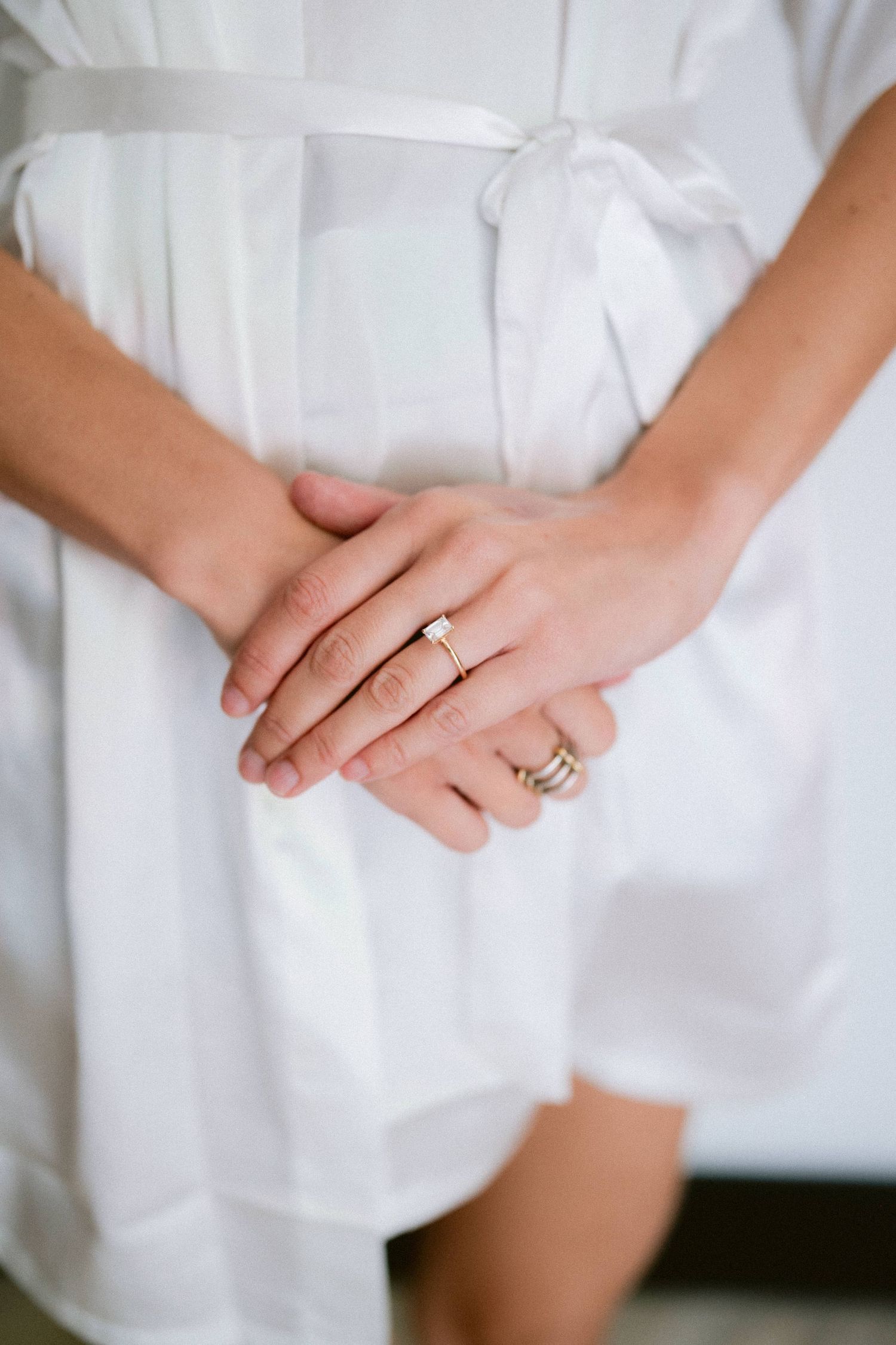 Close-up of bride's hands clasped together wearing a diamond engagement ring and white silk bridal robe.