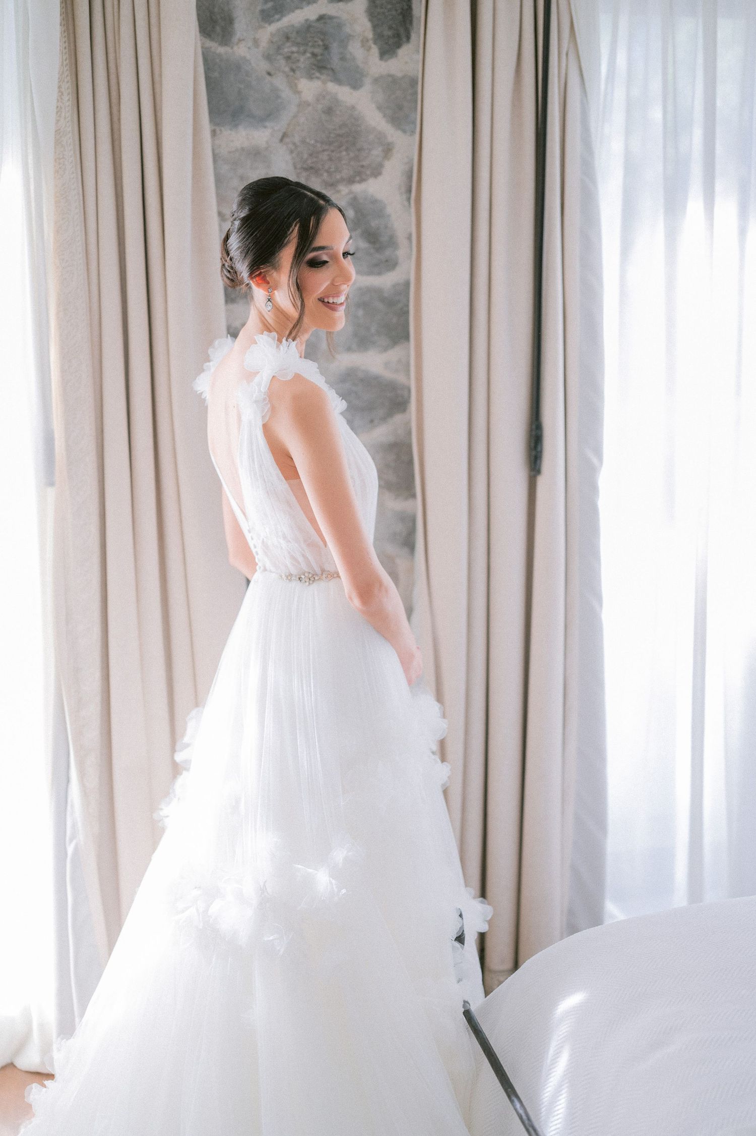 Smiling bride in layered tulle ball gown with feather details stands by bright window.