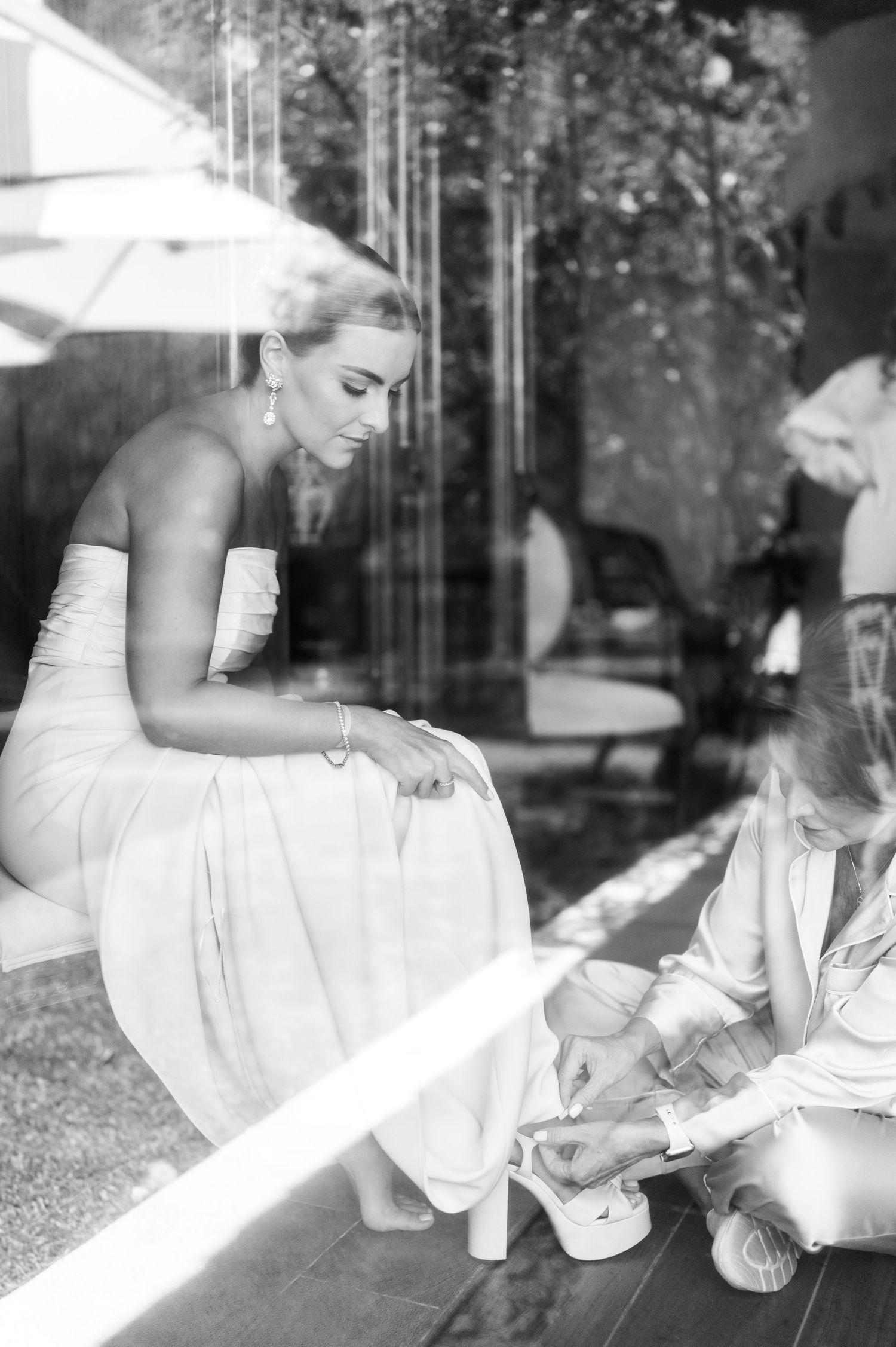 Black and white photo of bride in strapless gown having her shoes fastened through a glass window.