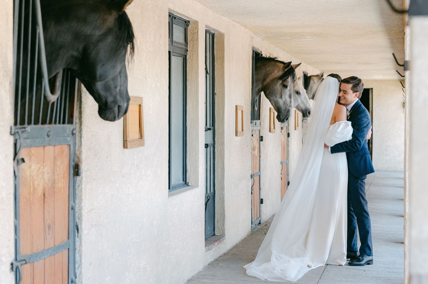 Bride and groom sharing a romantic kiss beside horse stables with horses looking on.