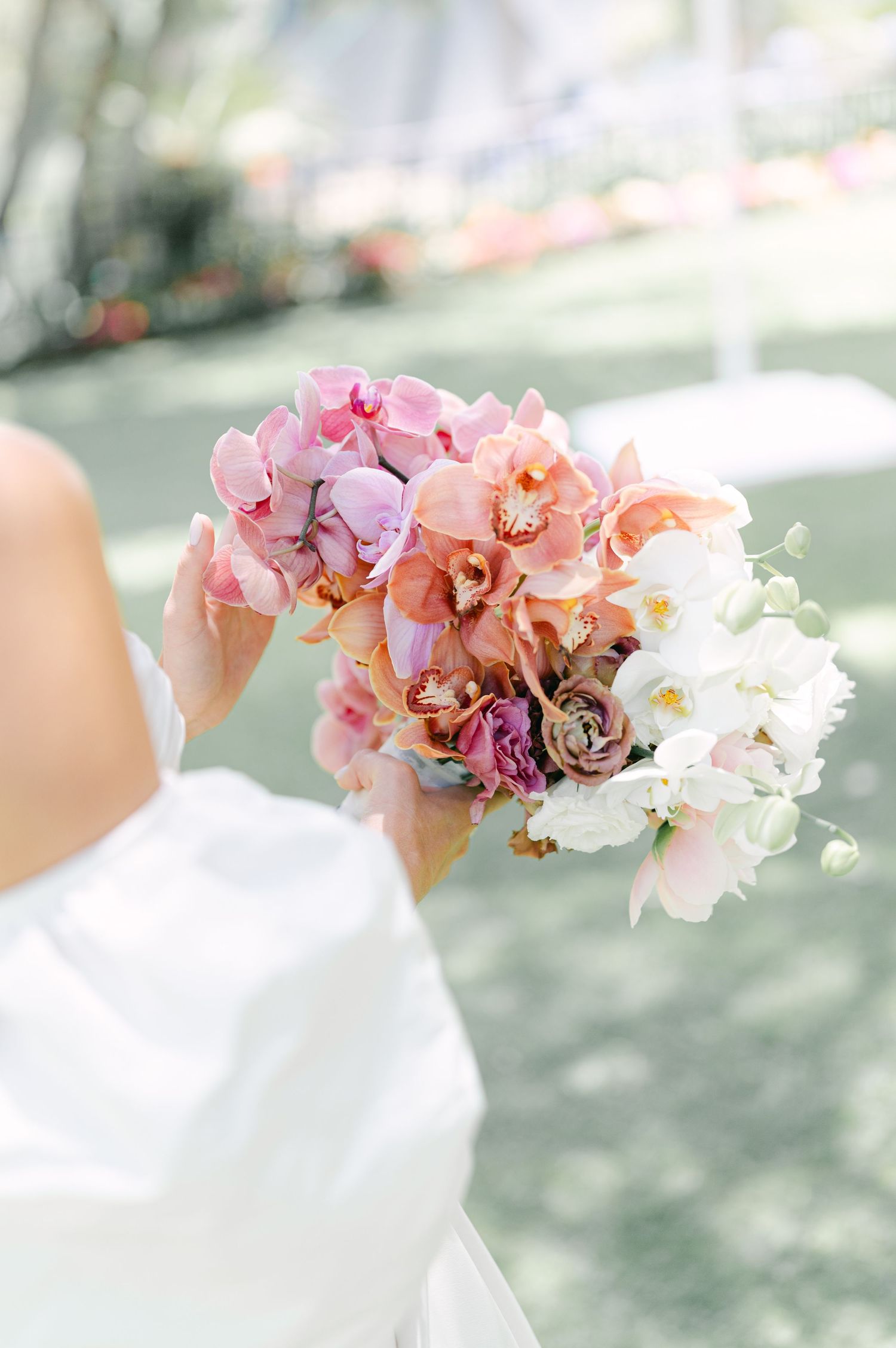 Bride holds vibrant bouquet of pink, peach, and white orchids with mauve ranunculus in sunlit outdoor setting.