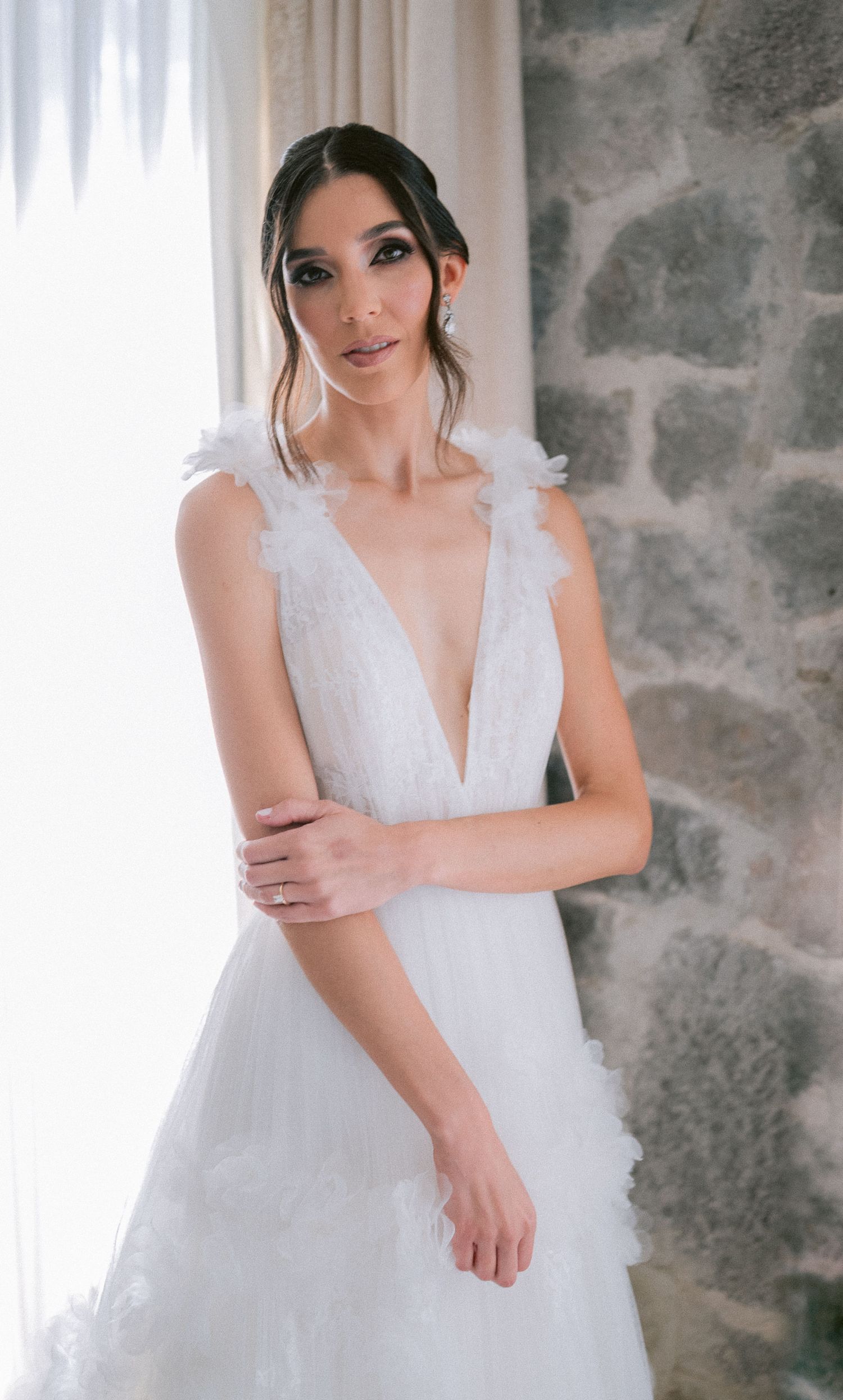Bride in deep V-neck white wedding gown with feather straps poses elegantly against stone wall.