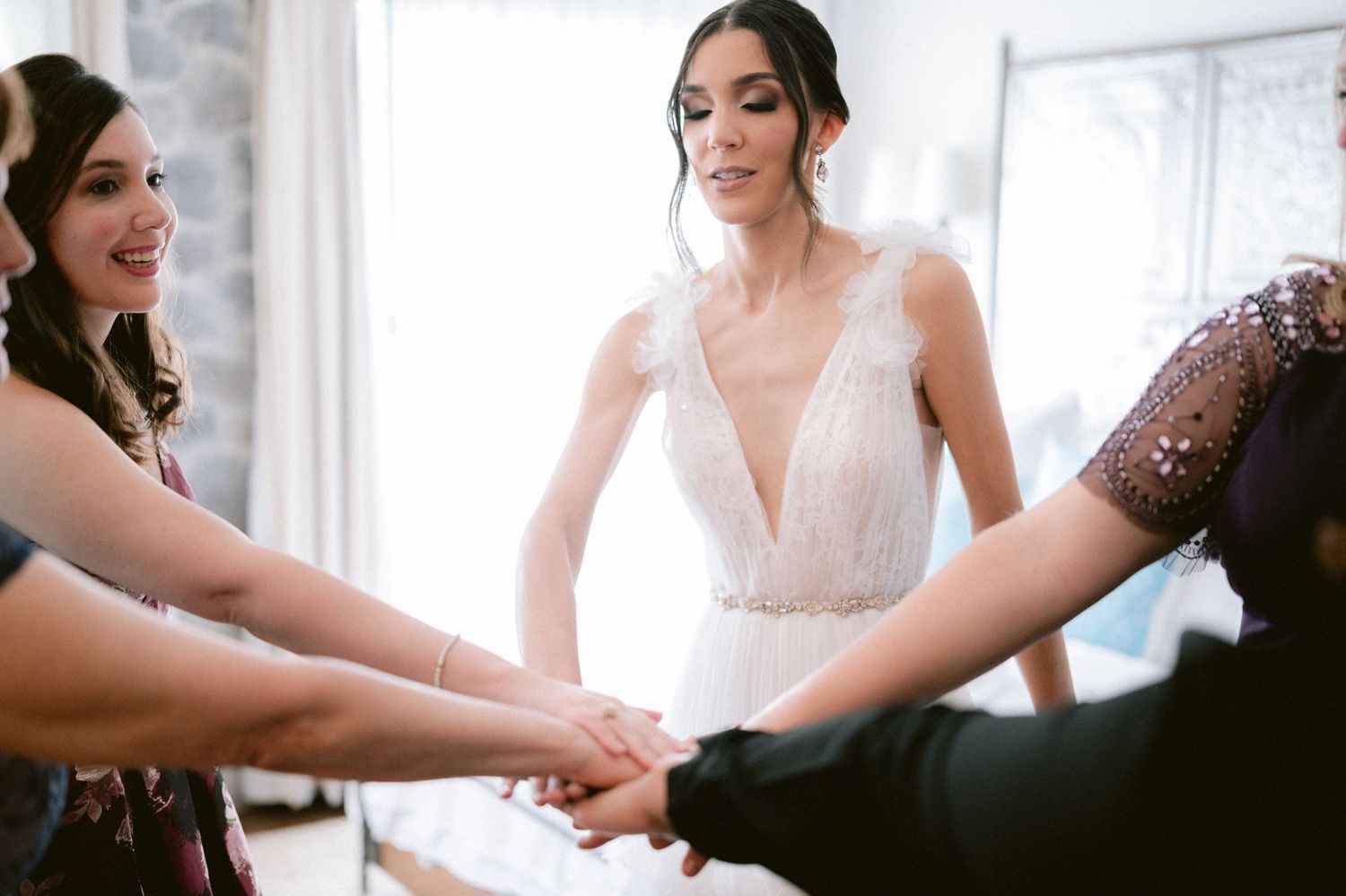 Bride in white feathered wedding dress joins hands with bridesmaids in a circle of support.