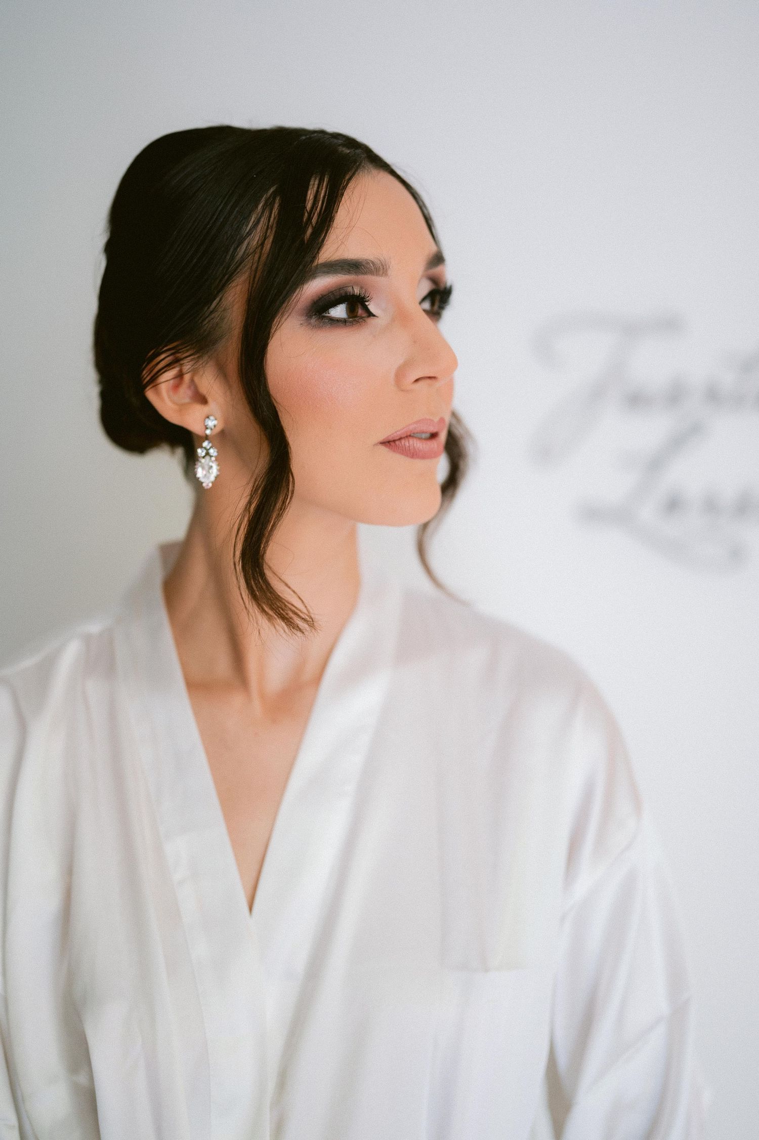Elegant bride in profile with dark upswept hair, dramatic eye makeup, and crystal earrings in white robe.