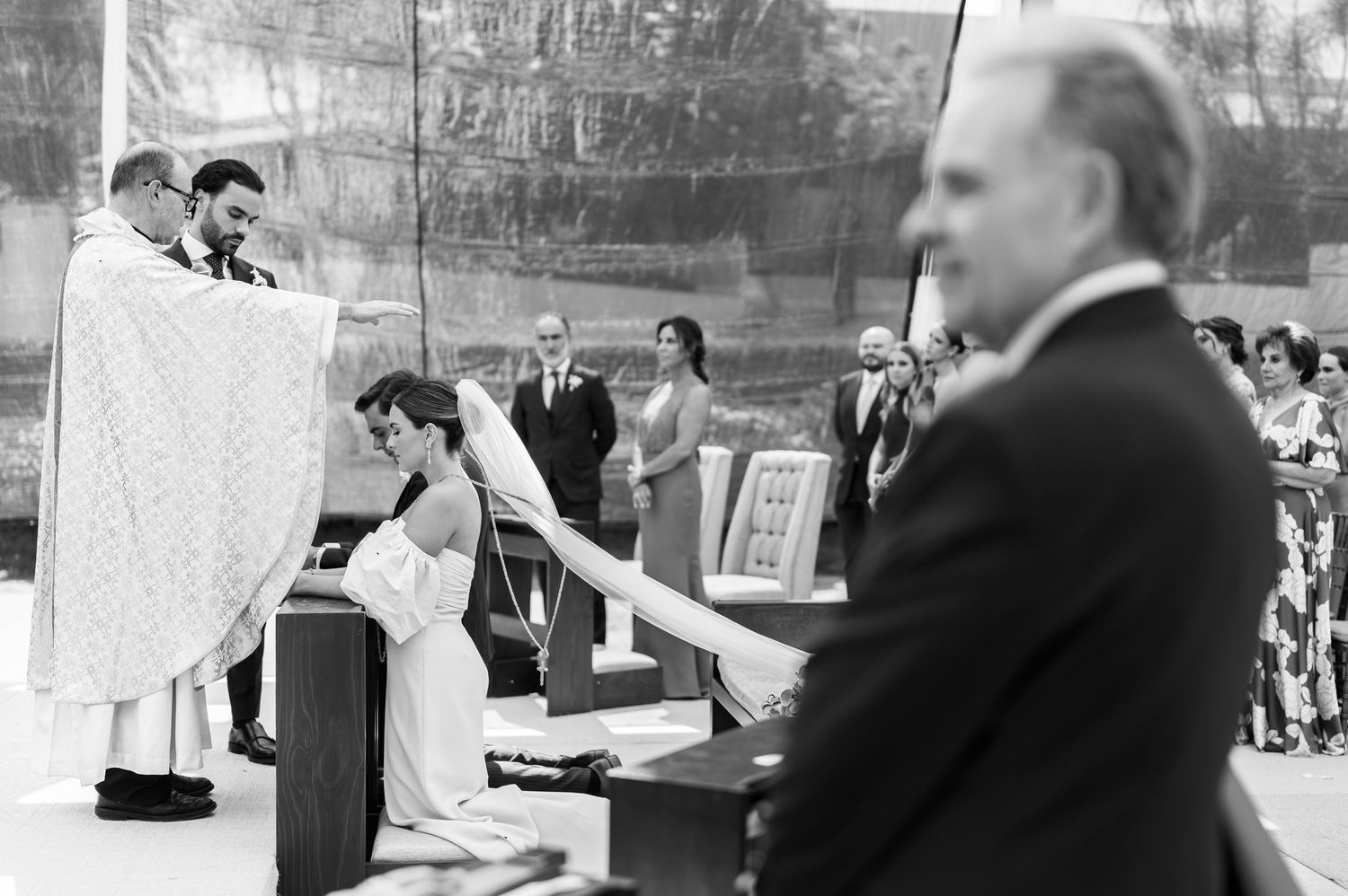 Black and white photo of couple kneeling before priest during formal outdoor wedding ceremony.