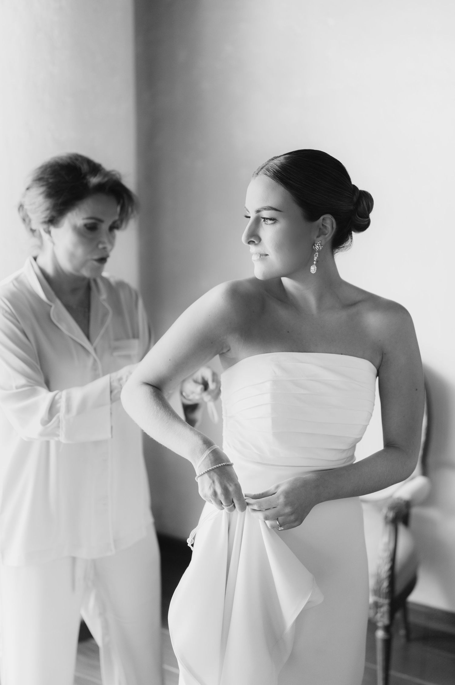 Black and white photo of mother helping bride into a strapless white wedding gown.
