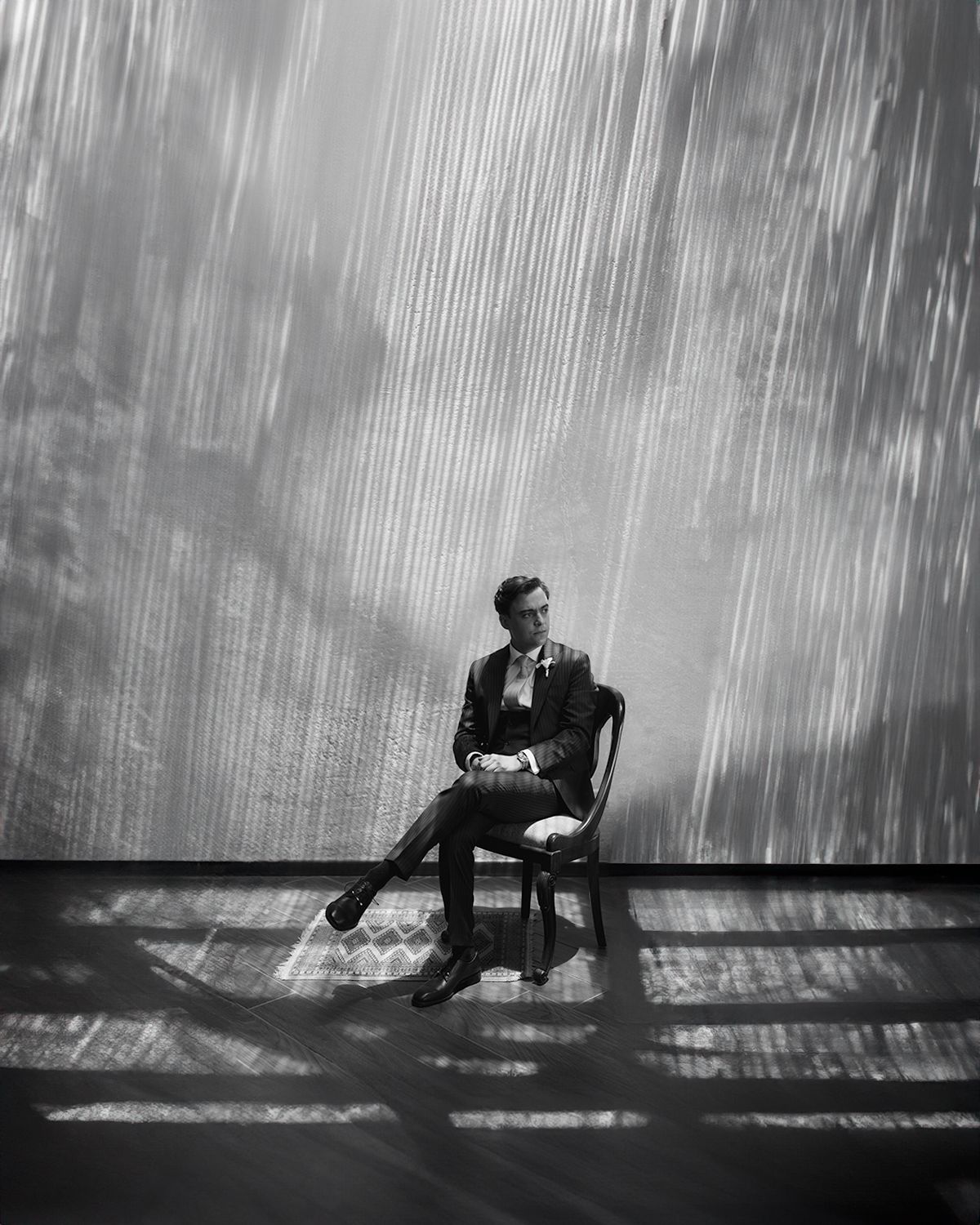 Elegant groom in a tailored suit seated in a chair against a dramatic light-streaked black and white backdrop.