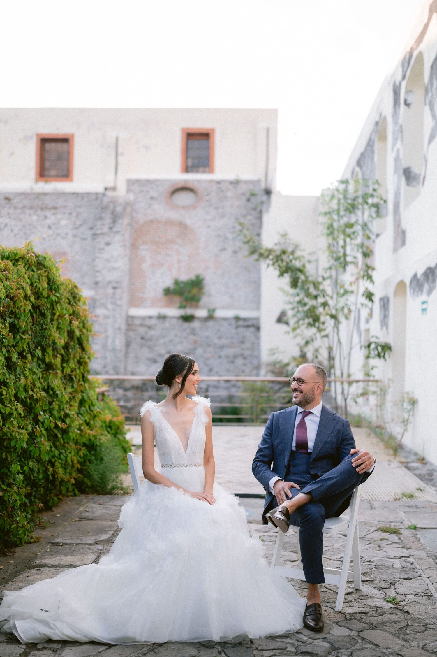Bride and groom sit casually in courtyard of rustic stone wedding venue smiling at each other.