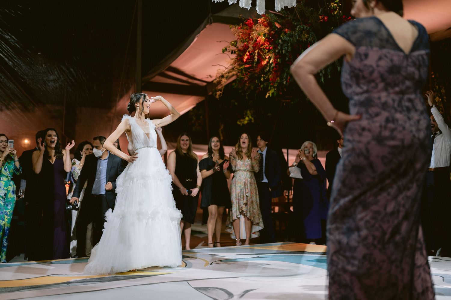 Bride in white ruffled gown dances joyfully on illuminated dance floor surrounded by cheering guests.