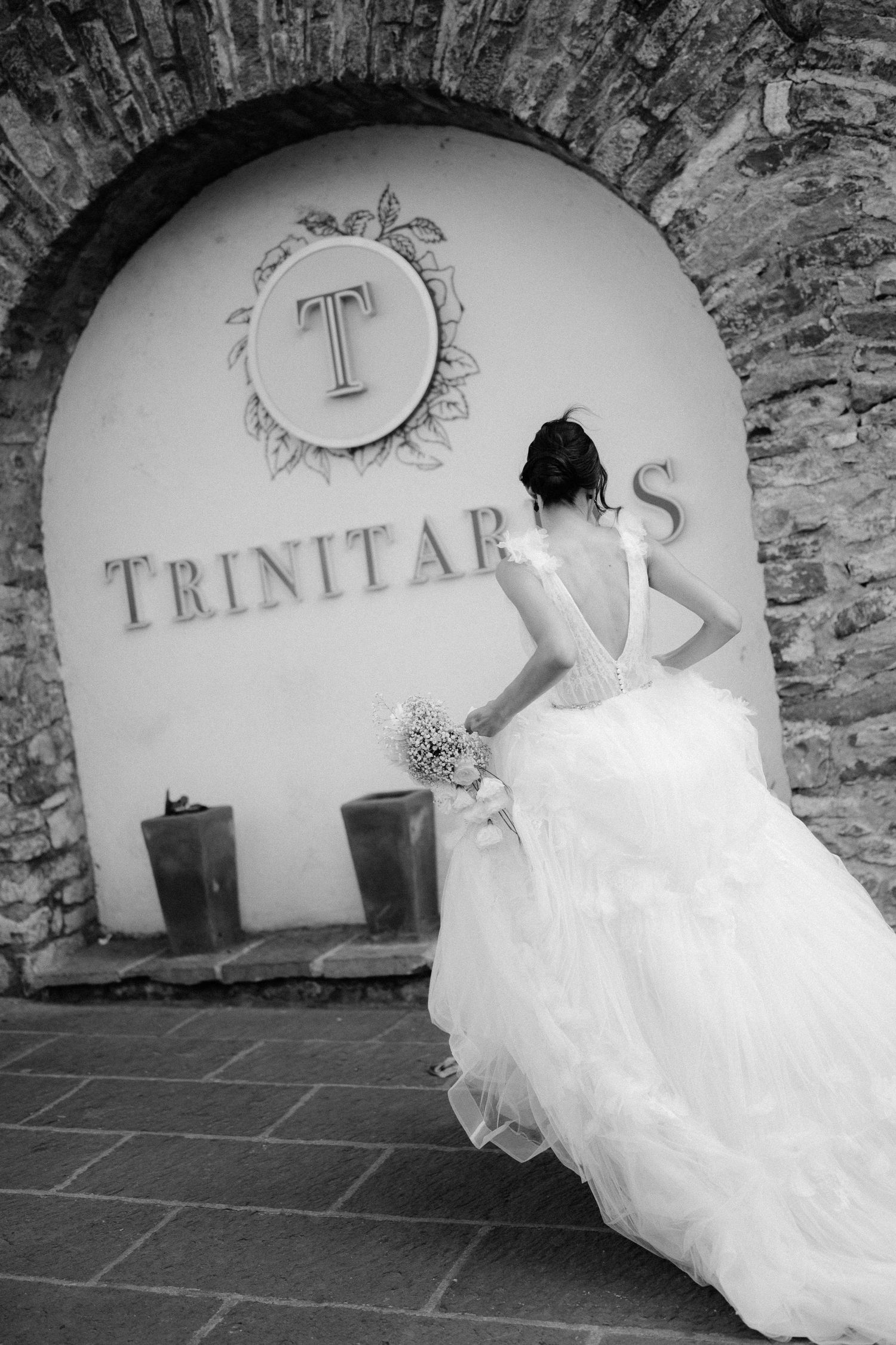 Black and white photo of bride in backless gown at Trinitarias stone arch venue entrance.