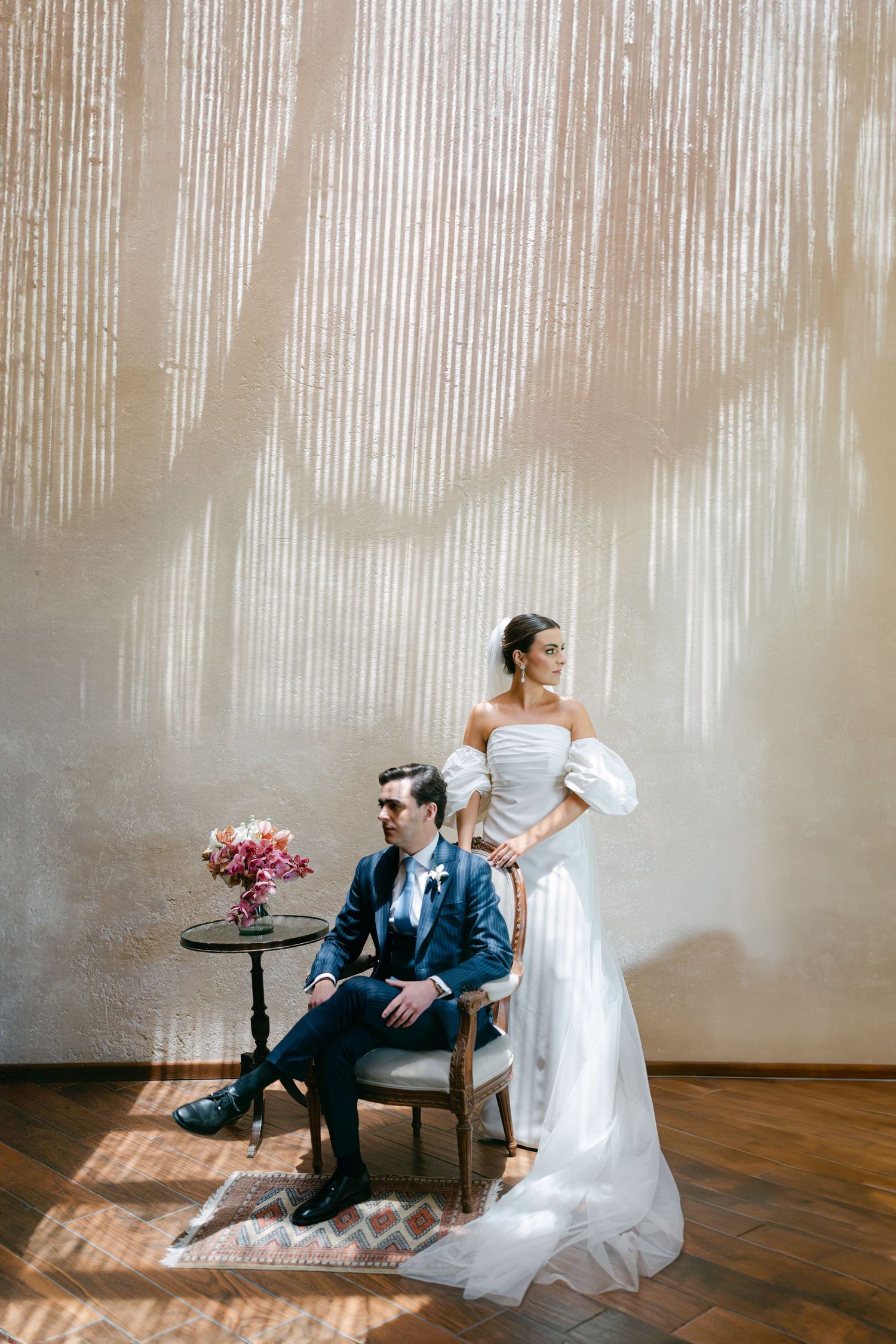 Bride in off-shoulder white gown stands behind groom seated in a vintage chair against a sunlit textured wall.