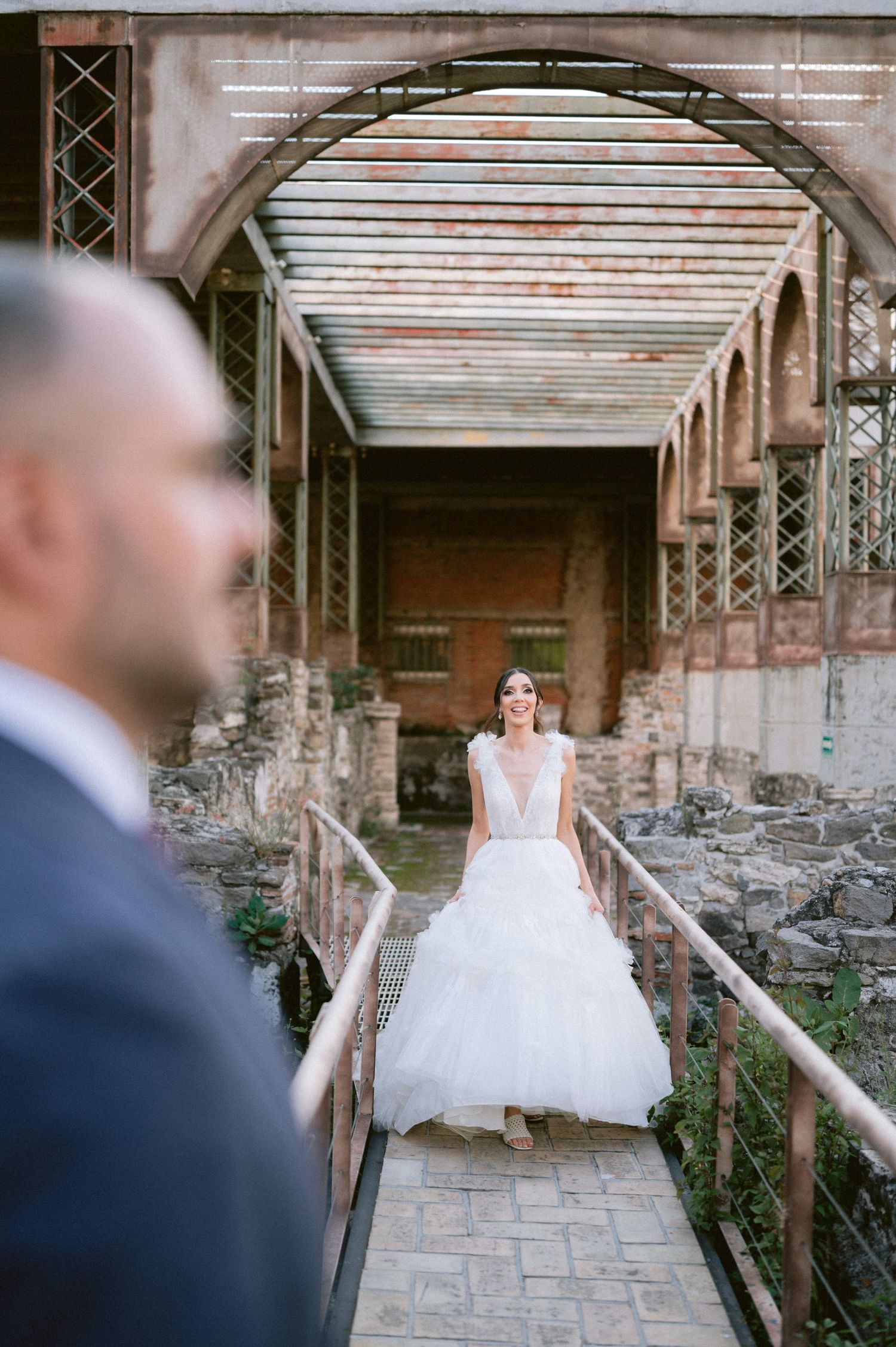 Bride in deep V-neck tulle gown walks toward groom on rustic bridge in historic industrial venue setting.