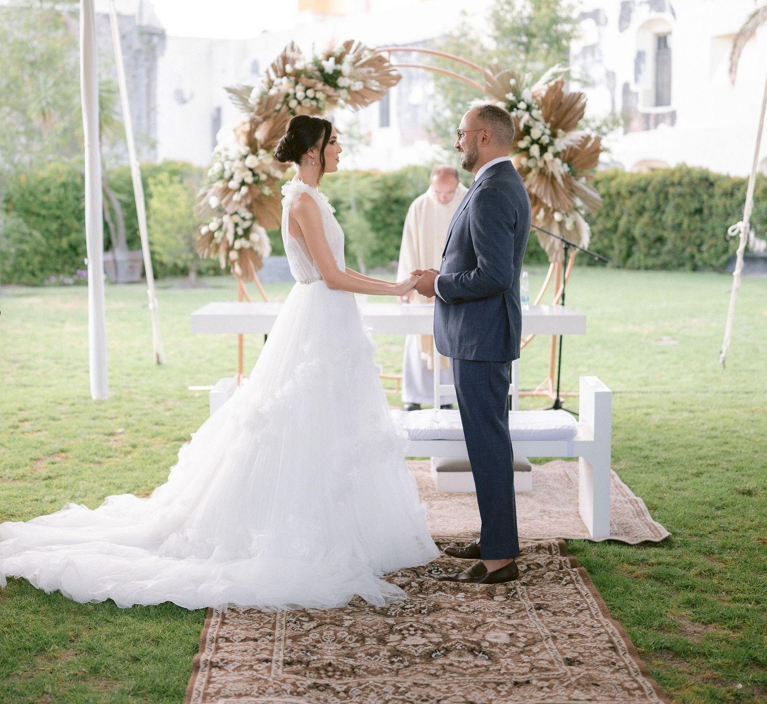 Bride and groom exchange vows outdoors at elegant boho wedding ceremony with pampas grass floral arch.