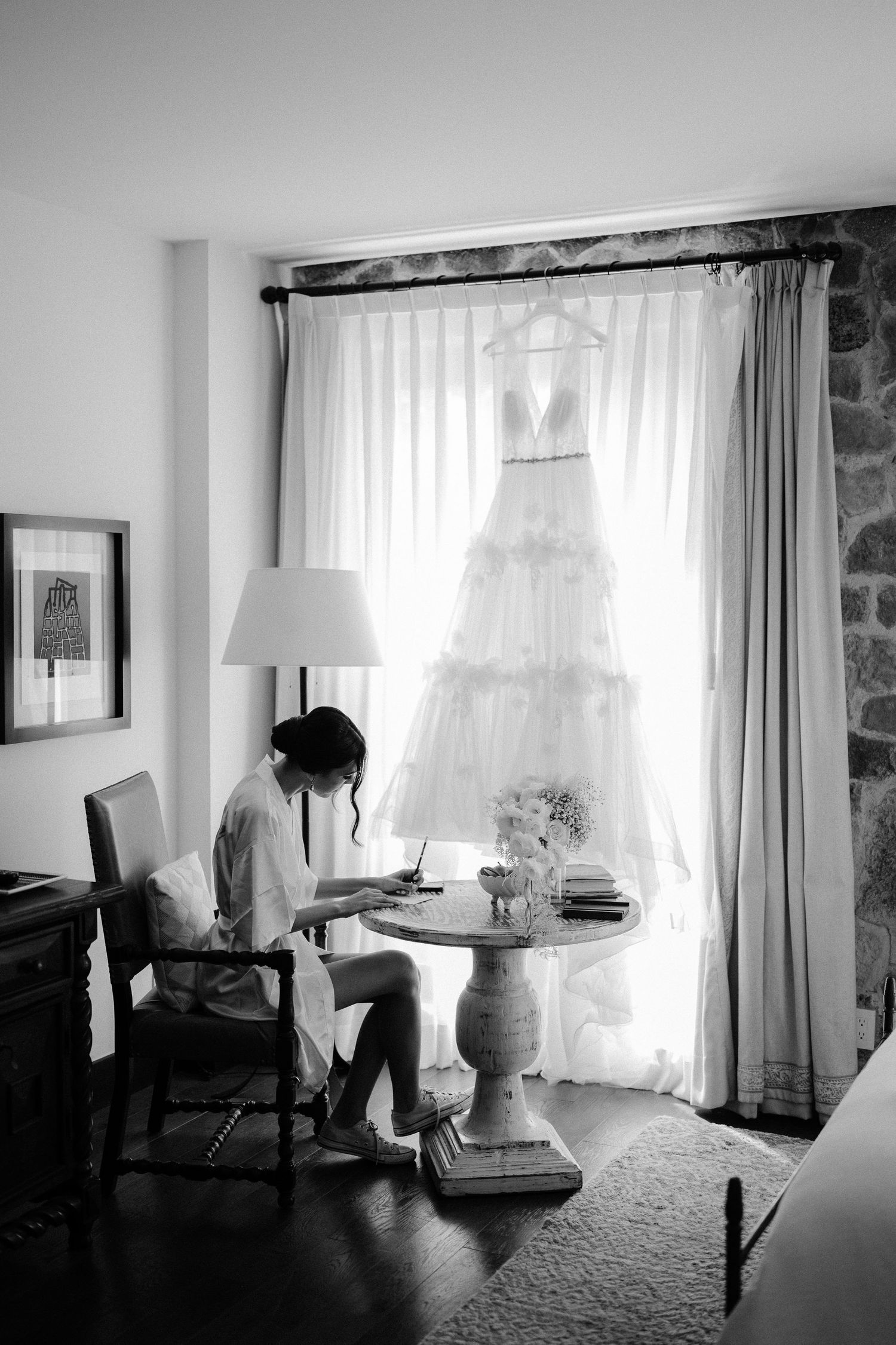 Black and white photo of bride writing at table with wedding gown hanging in window behind her.