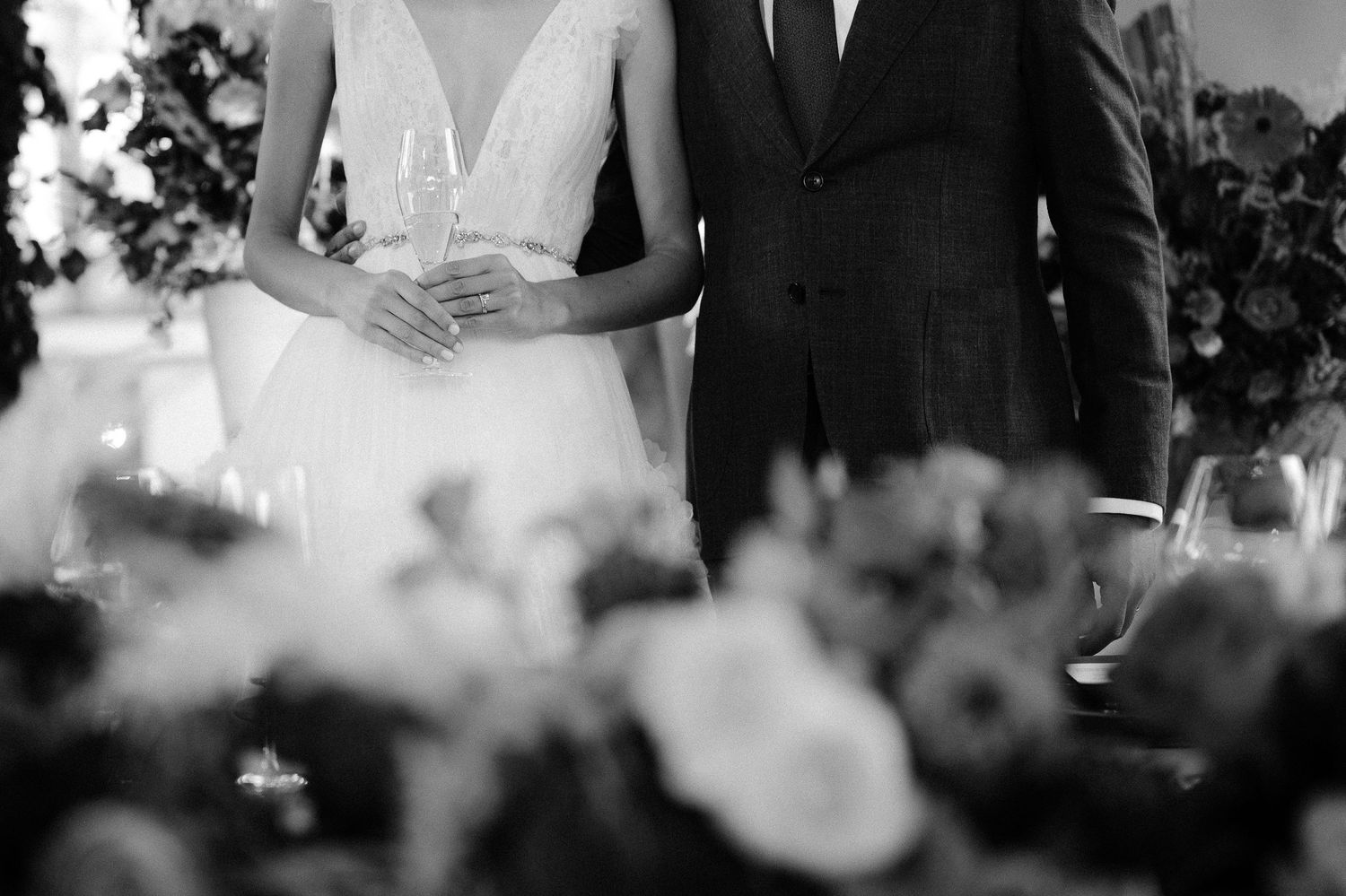 Black and white photo of bride and groom standing together at a floral-decorated wedding reception table.